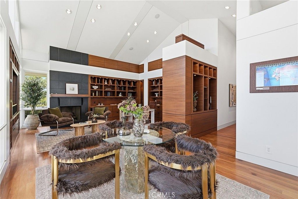 This spacious, open-concept living area features a vaulted ceiling and warm wood flooring throughout. The room is anchored by a dark-toned fireplace surround and built-in shelving, while a circular glass-topped dining table with textured chairs sits in the foreground. The perspective captures a bright, airy atmosphere with a seamless transition between the seating area and the adjacent hallway.