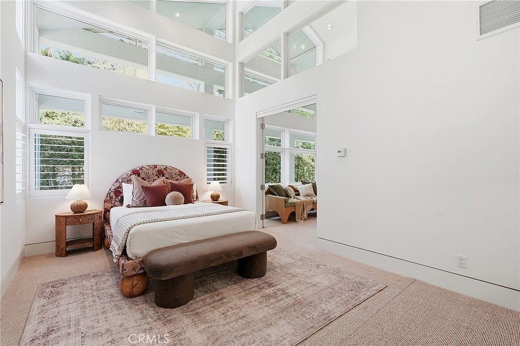 This spacious primary bedroom features a dramatic double-height ceiling with multiple tiers of large windows that flood the room with natural light. The space is anchored by a bed with a distinctive floral-patterned headboard, complemented by a neutral-toned rug and a dark, low-profile bench at the foot of the bed. A doorway leads into an adjacent sun-filled sitting area, creating an open and airy atmosphere with a modern, architectural aesthetic.
