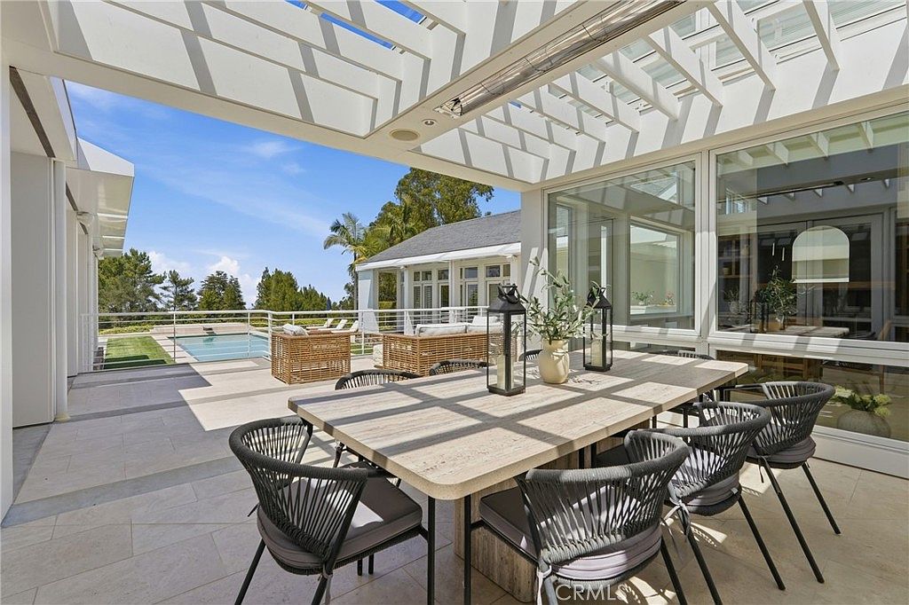 This expansive covered patio features a modern dining set with woven chairs and a light-toned table, perfect for outdoor entertaining. The space is sheltered by a geometric pergola and opens directly onto a sun-drenched pool deck with additional lounge seating. Large glass panels provide a seamless transition between the indoor and outdoor living areas, offering a bright and airy atmosphere with views of the surrounding landscape.