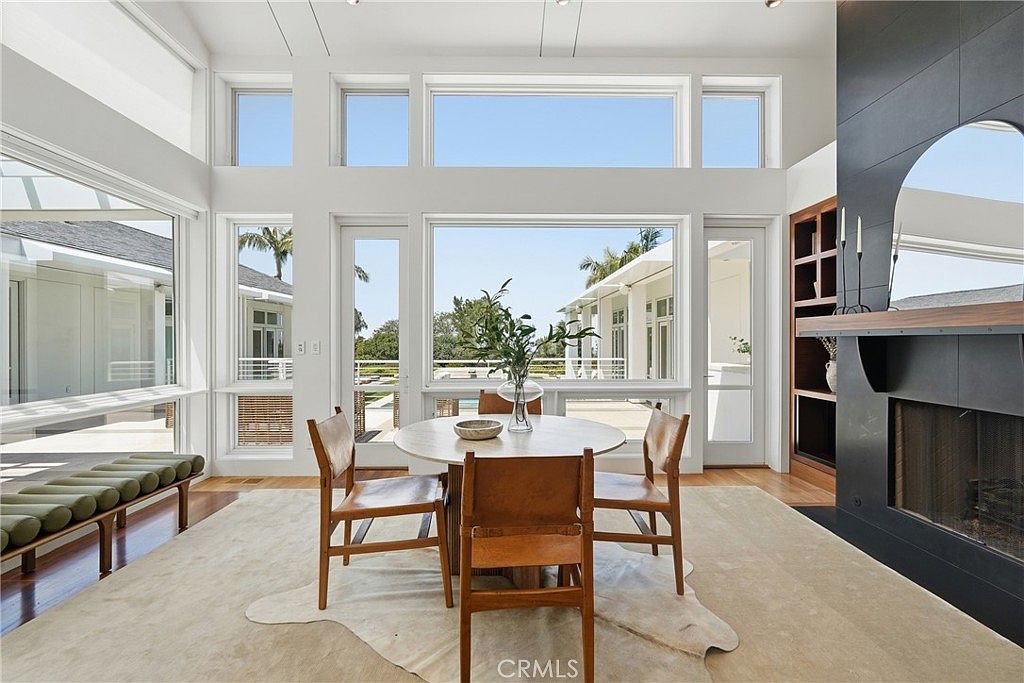 This bright dining area features a round wooden table surrounded by three mid-century modern style chairs, set atop a light-colored hide rug. The space is defined by expansive windows that offer a clear view of the outdoor patio and pool area, creating a seamless indoor-outdoor connection. To the right, a dark, full-height fireplace surround adds a dramatic contrast to the light, airy atmosphere of the room.