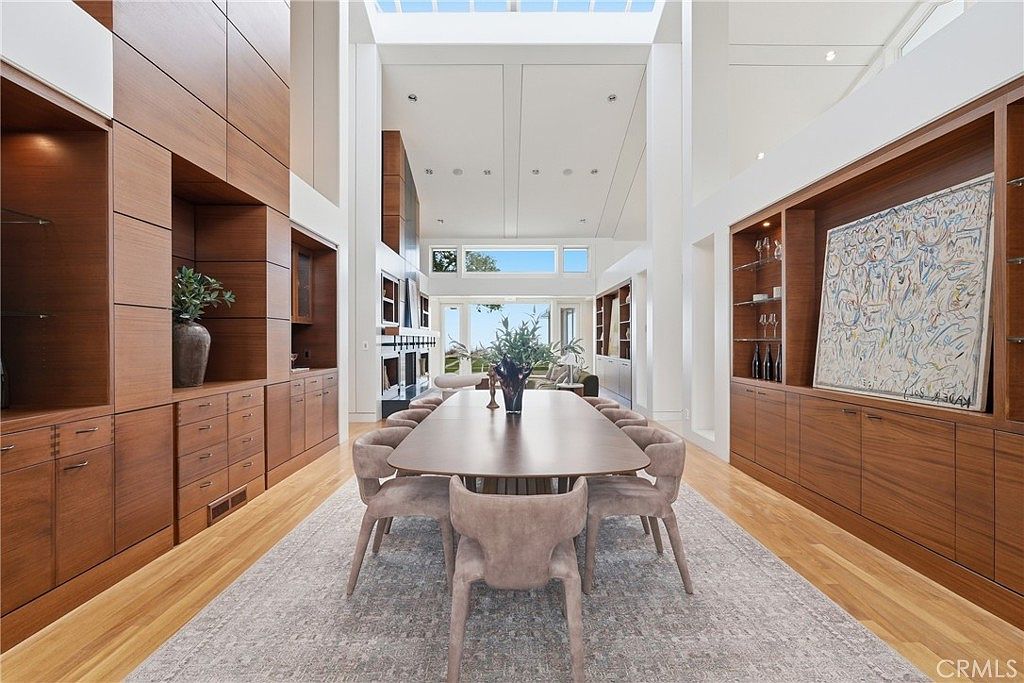 This spacious dining room features a long, dark wood table surrounded by ten upholstered chairs, centered on a large patterned area rug. The room is flanked by extensive custom wood cabinetry with integrated shelving and storage, creating a sophisticated and symmetrical aesthetic. A dramatic, high-vaulted ceiling with a skylight provides abundant natural light, while the open floor plan leads the eye toward a bright living area and expansive windows overlooking the outdoors.