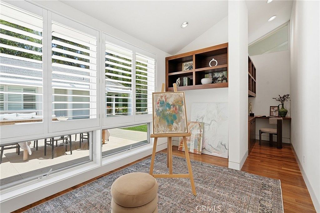 This bright, multi-functional room serves as an art studio and home office, featuring warm wood flooring and a large area rug. A wooden easel stands prominently in the center, while built-in shelving and a dedicated desk nook provide organized storage and workspace. Large windows with white plantation shutters allow ample natural light to fill the space, creating an inviting and creative atmosphere.