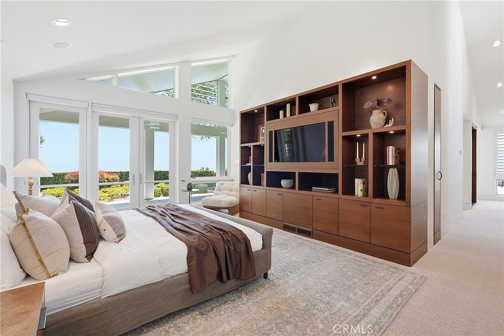 This spacious primary bedroom features a vaulted ceiling and large windows that provide abundant natural light and views of the outdoor greenery. A prominent, custom-built wood media unit with integrated shelving and cabinetry serves as a focal point, while a plush bed with neutral-toned bedding anchors the room. The space is finished with a soft, patterned area rug and a clean, minimalist aesthetic that creates a serene and inviting atmosphere.