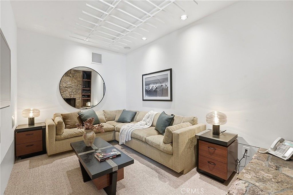 This cozy living room features a large, neutral-toned sectional sofa arranged around a modern, dark-toned coffee table. A circular mirror on the wall reflects a stone-clad wall, adding depth and texture to the space, while two matching table lamps provide warm ambient lighting. The room is finished with a light-colored carpet and a minimalist ceiling light fixture, creating a comfortable and inviting atmosphere.