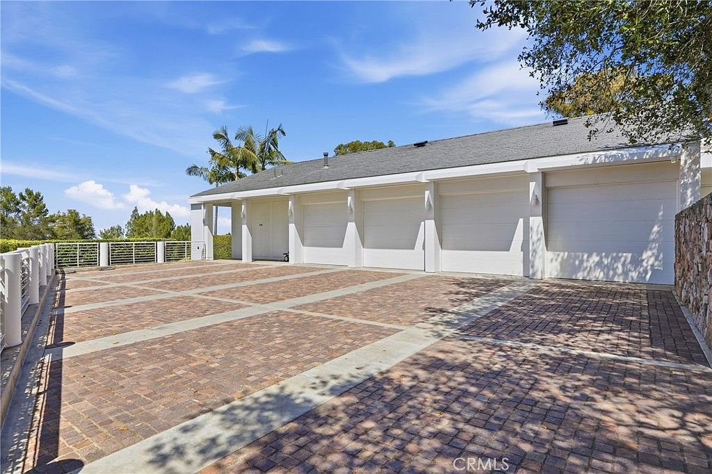 This image showcases a spacious, multi-car garage exterior featuring a large, paved driveway with a geometric pattern. The structure has a clean, white facade with multiple garage doors and a simple, low-pitched roofline. The scene is captured from a low angle, emphasizing the expansive driveway and the bright, sunny atmosphere of the property.