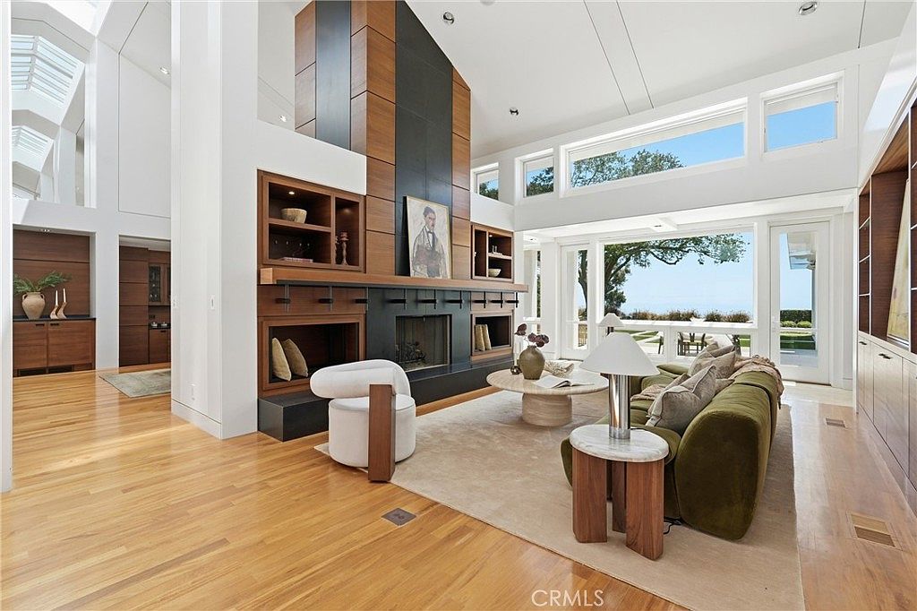 This spacious living room features a dramatic, tall fireplace surround with a dark, modern finish that serves as the central focal point. The room is bathed in natural light from large windows and clerestory windows, highlighting the light wood flooring and neutral color palette. A plush olive-green sofa and a circular coffee table create a comfortable seating area, while the open-concept design provides a seamless transition to the outdoor view.