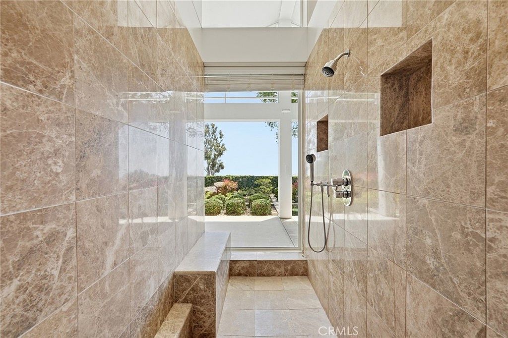 This luxurious walk-in shower features floor-to-ceiling stone tile walls and a built-in stone bench. A glass door opens directly to an outdoor garden area, creating a seamless indoor-outdoor transition. The shower is equipped with both a fixed overhead showerhead and a handheld sprayer, offering a spa-like experience.