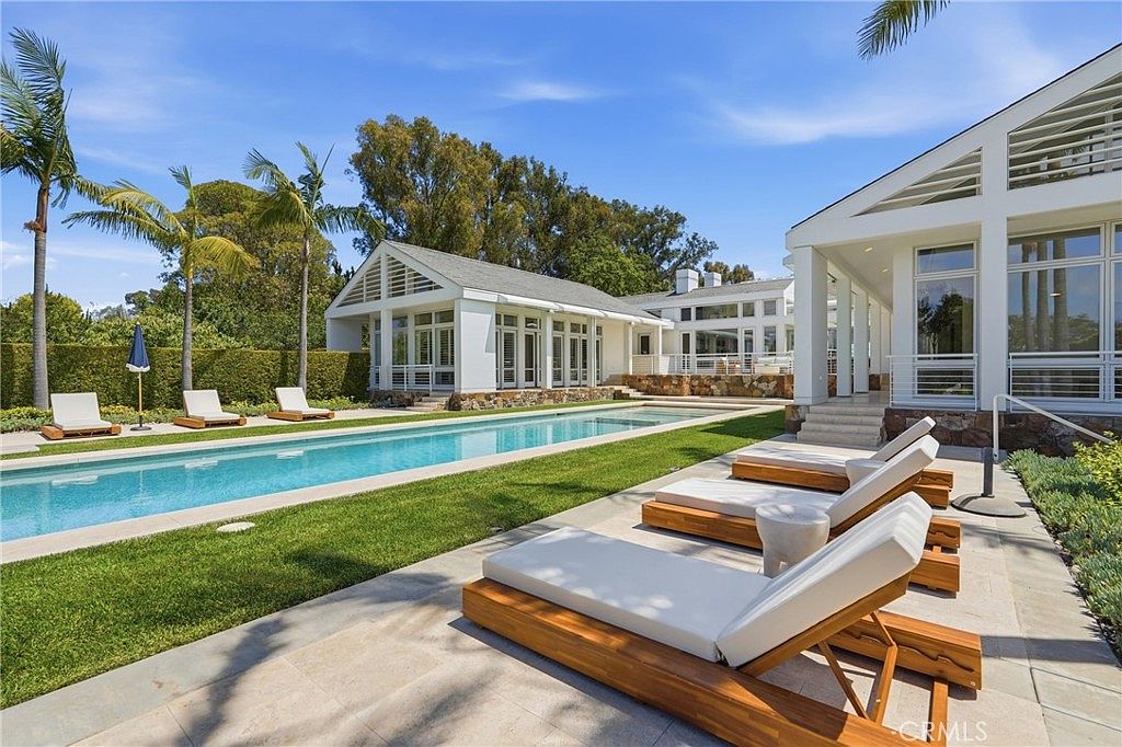 This bright, sun-drenched backyard features a long, rectangular swimming pool as the central focal point, surrounded by a stone patio and manicured lawn. Several wooden lounge chairs with white cushions are arranged neatly on the patio, inviting relaxation in this serene outdoor space. The property includes a white, modern-style structure with large windows and glass doors that seamlessly connect the indoor and outdoor living areas.
