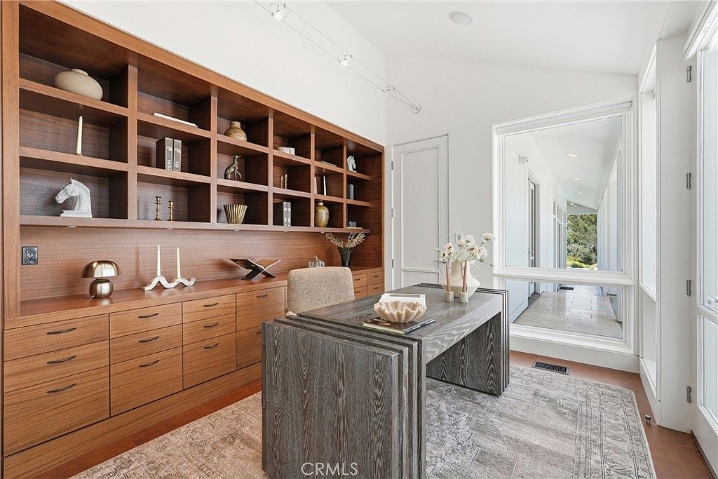 This sophisticated home office features extensive built-in cabinetry with open shelving and lower drawers in a warm wood finish. A textured, dark-toned desk sits centrally on a patterned area rug, complemented by a neutral-toned office chair. Large windows provide ample natural light and a view of the outdoor area, creating a bright and professional workspace.