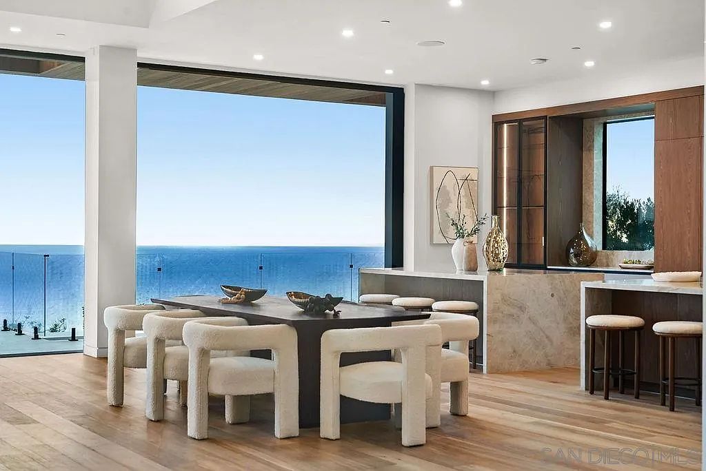 This sophisticated dining area features a dark rectangular table surrounded by six cream-colored, textured chairs, positioned to take full advantage of the expansive ocean view through large glass walls. The open-concept space transitions seamlessly into a kitchen area with a stone-topped island and bar seating, all set against light wood flooring. The room is bathed in natural light, creating an airy and luxurious atmosphere that emphasizes the connection between the interior living space and the coastal landscape.