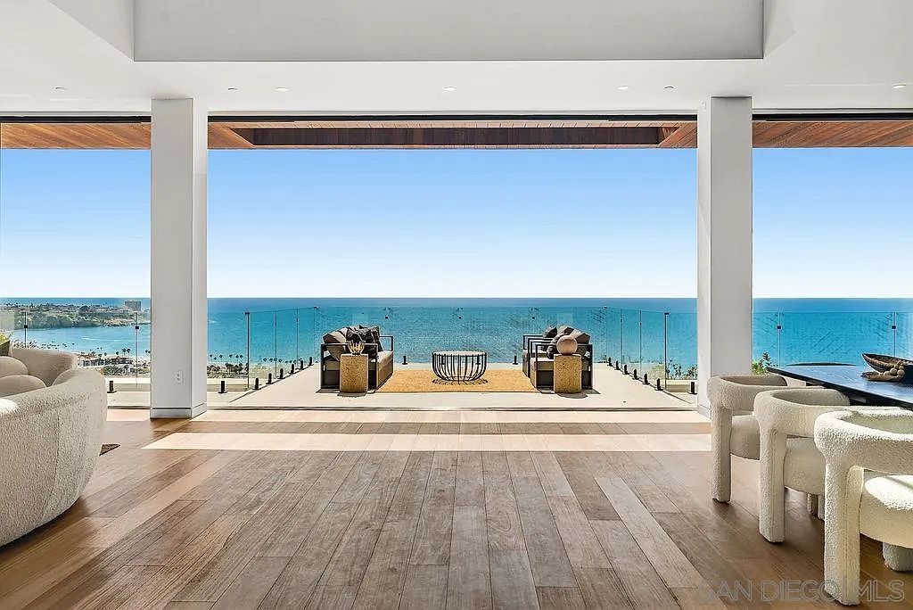This expansive living area features wide-plank wood flooring and a seamless transition to an outdoor terrace through massive, floor-to-ceiling glass walls. The interior is furnished with plush, light-toned seating, while the exterior deck offers a panoramic, unobstructed view of the ocean. The design emphasizes an open-concept layout that blurs the line between indoor comfort and the coastal landscape.