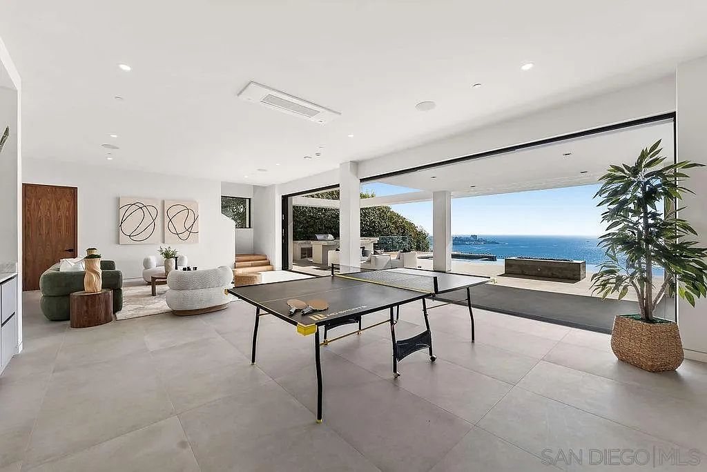 This expansive, open-concept living area features a modern aesthetic with large-format light-colored tile flooring and a seamless transition to an outdoor patio through wide-opening glass doors. The space is furnished with a comfortable sofa and lounge chairs, centered around a ping-pong table, while a large potted plant adds a touch of greenery. The room offers a stunning, unobstructed view of the ocean, creating a bright and airy atmosphere perfect for indoor-outdoor entertaining.