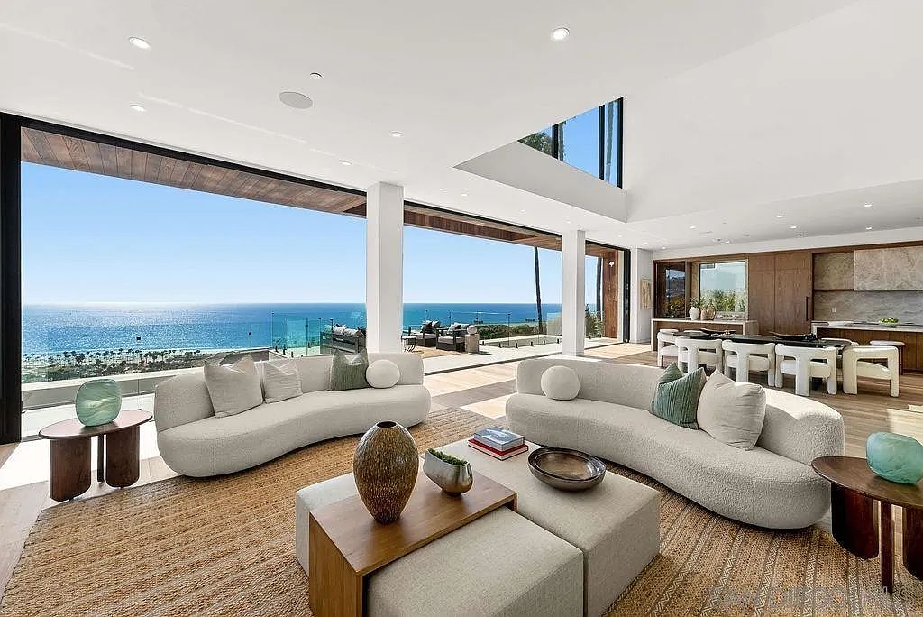 This expansive, open-concept living area features a modern aesthetic with two curved, light-toned sofas arranged around a central coffee table setup. The space is defined by massive floor-to-ceiling glass walls that provide an unobstructed, panoramic view of the ocean and coastline. The interior flows seamlessly into a kitchen area in the background, creating a bright, airy atmosphere characterized by neutral textures and a sophisticated, coastal-luxury design.