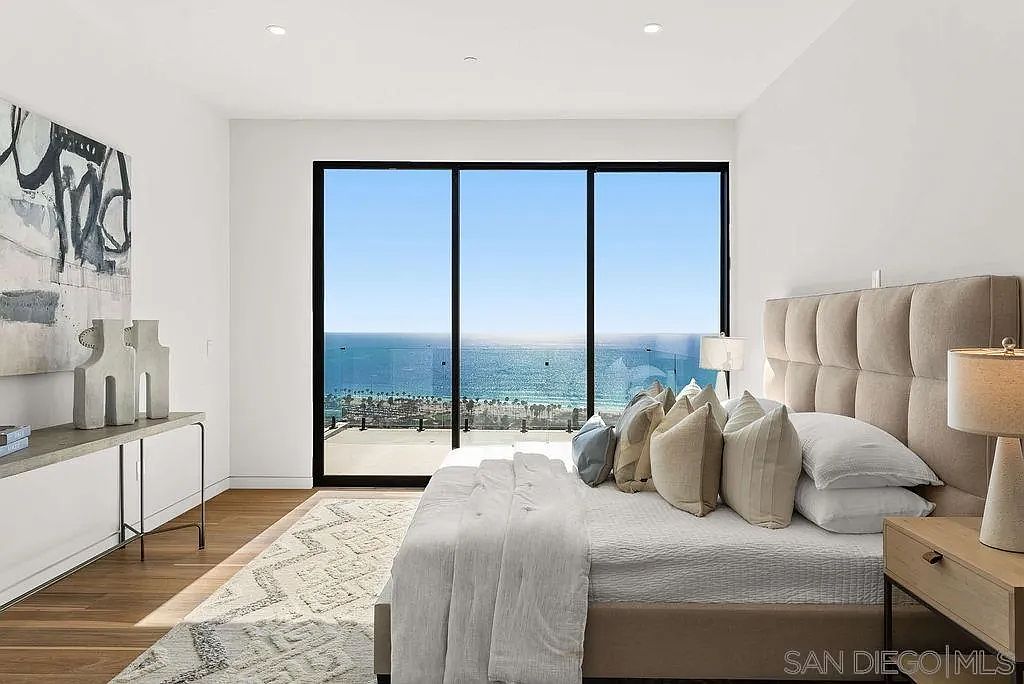 This primary bedroom features a neutral, modern aesthetic with a large, tufted upholstered headboard as the focal point. The room is anchored by a patterned area rug over light wood flooring and includes a sleek console table against the left wall. A wall of floor-to-ceiling glass provides an expansive, unobstructed view of the ocean and coastline, creating a serene and luxurious atmosphere.