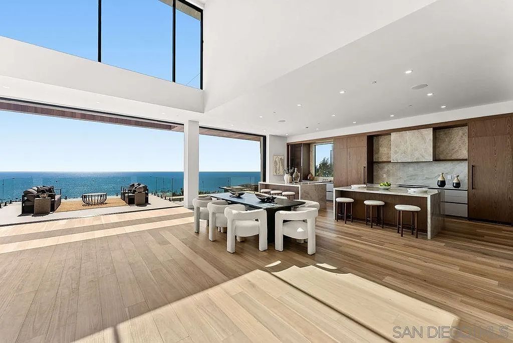 This expansive, open-concept living and dining area features light wood flooring and a seamless transition to an outdoor deck through massive, wall-to-wall glass panels. The space includes a modern kitchen with a large island, a formal dining table, and a living area, all oriented toward a panoramic ocean view. The minimalist design emphasizes natural light and a clean, airy aesthetic that highlights the connection between the interior and the coastal landscape.