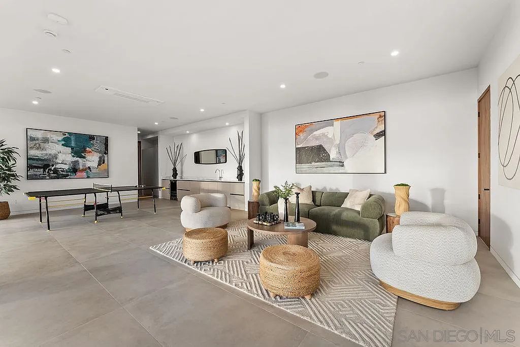 This spacious, open-concept living area features a contemporary design with large-format tile flooring and a neutral color palette. The room is furnished with a plush green sofa, two curved accent chairs, and a pair of woven ottomans centered on a geometric area rug. A ping-pong table is positioned in the background, while a sleek bar area with cabinetry and a mirror adds a sophisticated touch to the versatile space.
