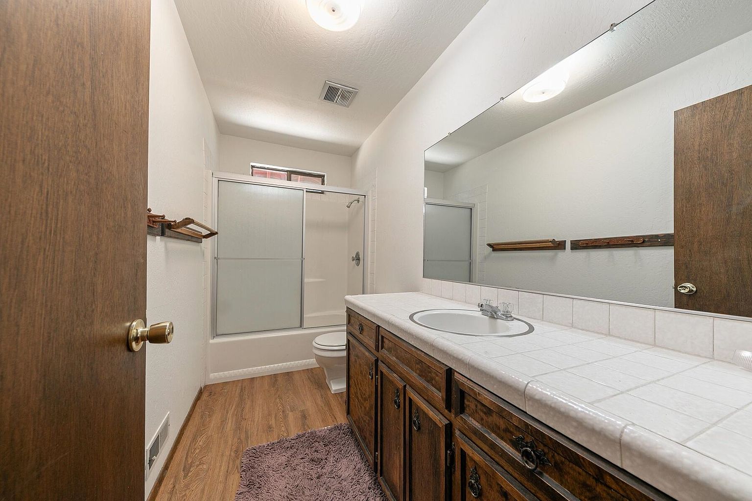 This is a bathroom featuring a vanity with dark wood cabinets and a white tiled countertop, a white sink, and a large mirror above the vanity. A shower/tub combination with sliding glass doors is visible in the background, along with a toilet. The flooring is wood-look laminate, and a small rug is on the floor.