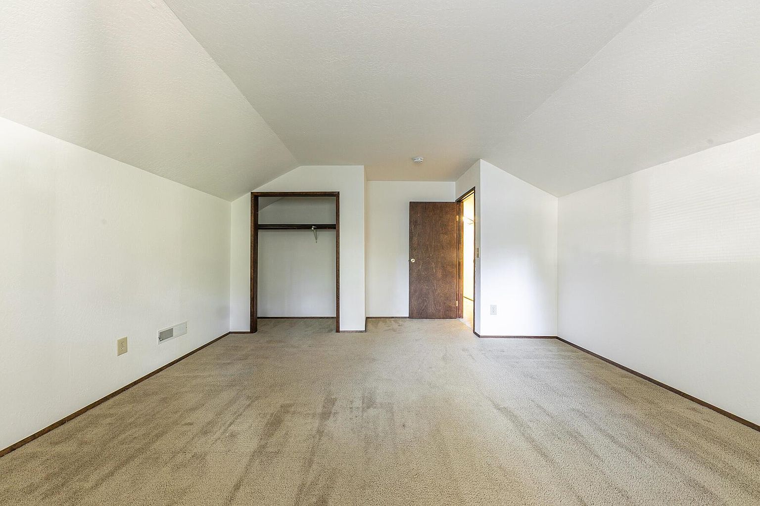 This is an interior shot of a bedroom, featuring neutral beige carpeting and white walls. The room includes a closet with a dark wood frame and a partially open wooden door. The space is well-lit and appears clean, offering a blank canvas for potential buyers.
