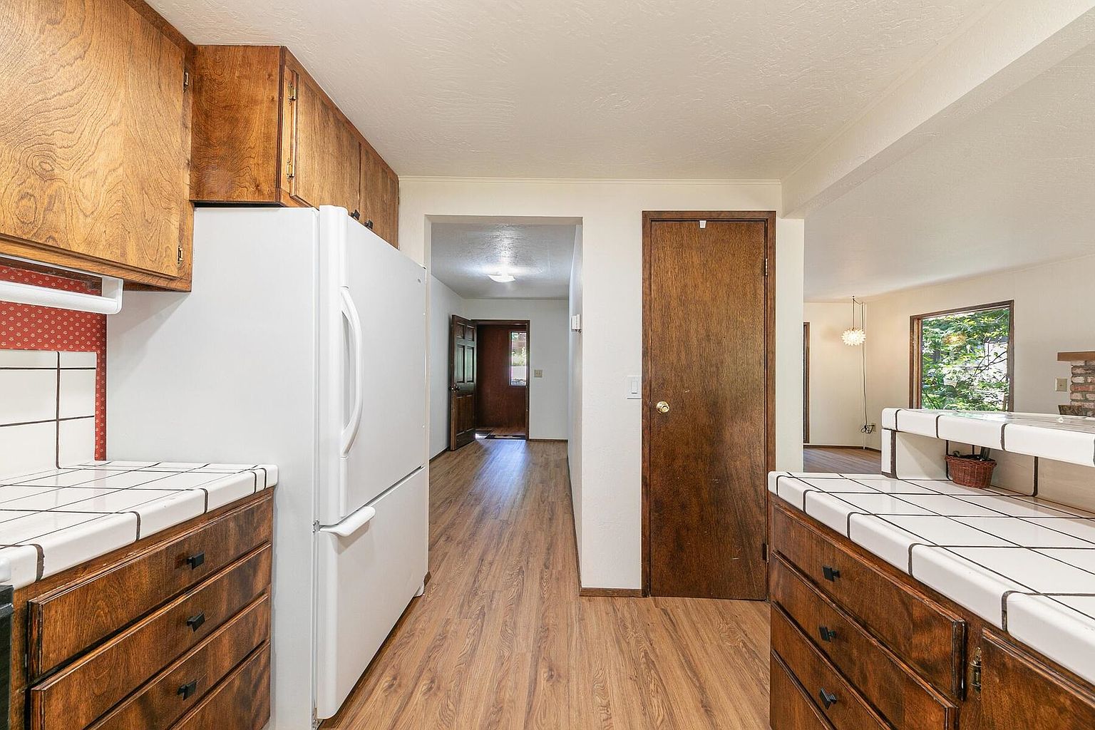 This kitchen features wooden cabinets and drawers with white tiled countertops. A white refrigerator stands against the cabinets, and the flooring is wood-look laminate. The view extends into a hallway and a glimpse of the living room, creating a sense of openness.