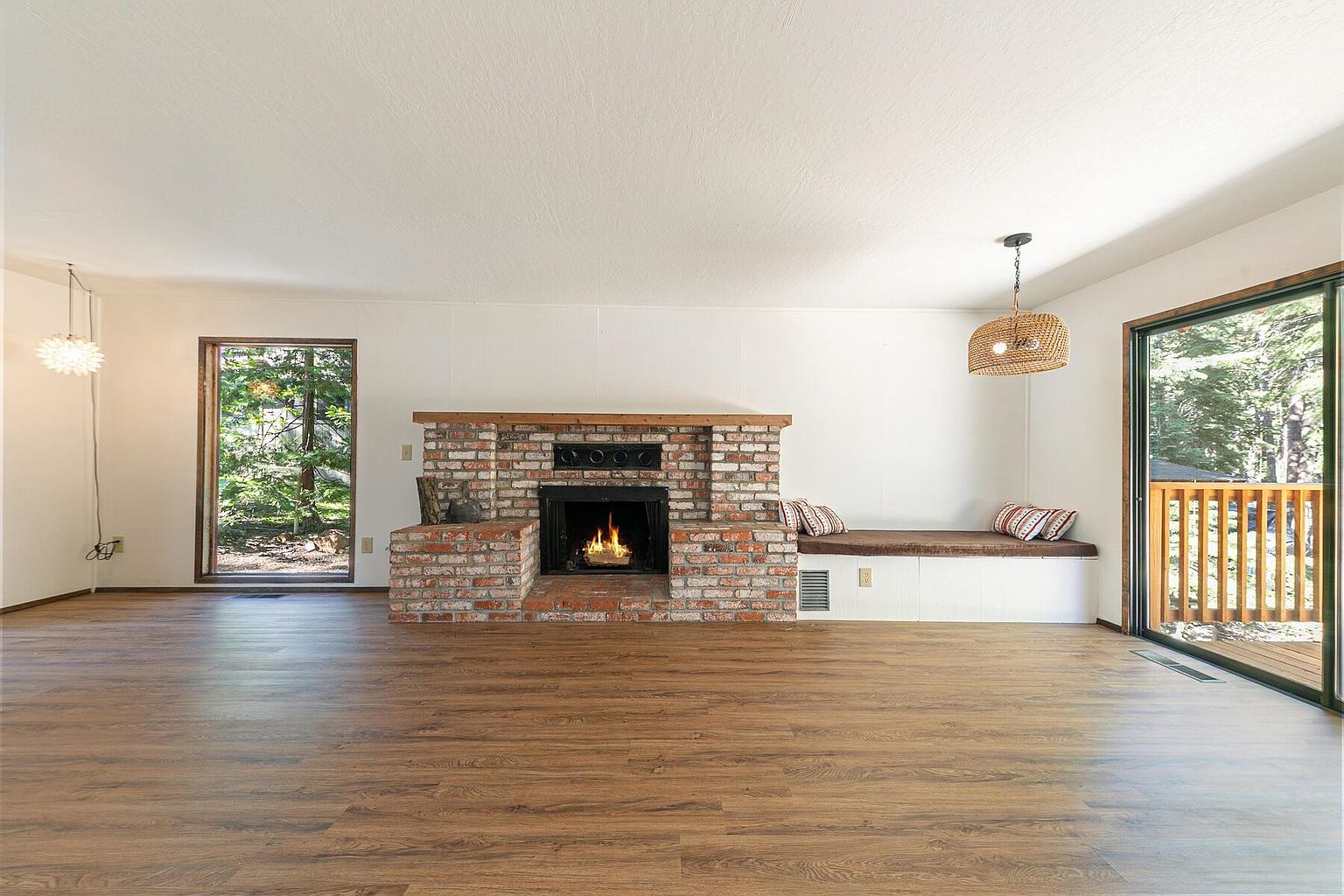 This is an interior shot of a living room featuring hardwood floors, a brick fireplace, and a built-in window seat. Natural light streams in through a large window and sliding glass door, highlighting the room's warm and inviting atmosphere. The room is spacious and appears well-maintained, offering a comfortable living space.