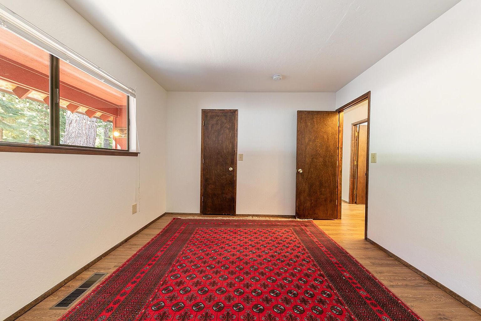 The image showcases a bedroom with a red patterned rug covering most of the wooden floor. The walls are painted in a neutral tone, and a window provides natural light. Two wooden doors are visible, one closed and one slightly ajar, suggesting access to other areas of the house.