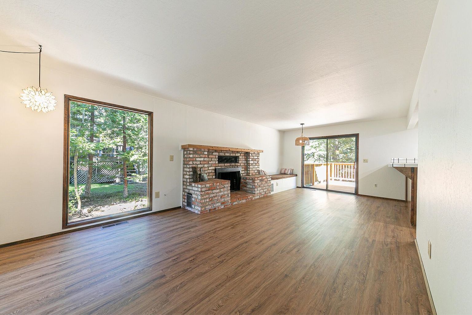 This is a bright and spacious living room featuring hardwood floors and white walls. A brick fireplace serves as a focal point, complemented by a large window offering natural light and views of the outdoors. Sliding glass doors lead to an outdoor deck, enhancing the indoor-outdoor flow.