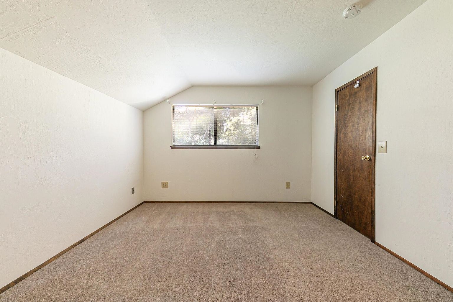 The image showcases a simple, carpeted bedroom with neutral-toned walls and a single window providing natural light. A dark wood door is visible on the right, and the room appears clean and uncluttered. The space has a basic, functional aesthetic, suitable for a guest bedroom or secondary bedroom.
