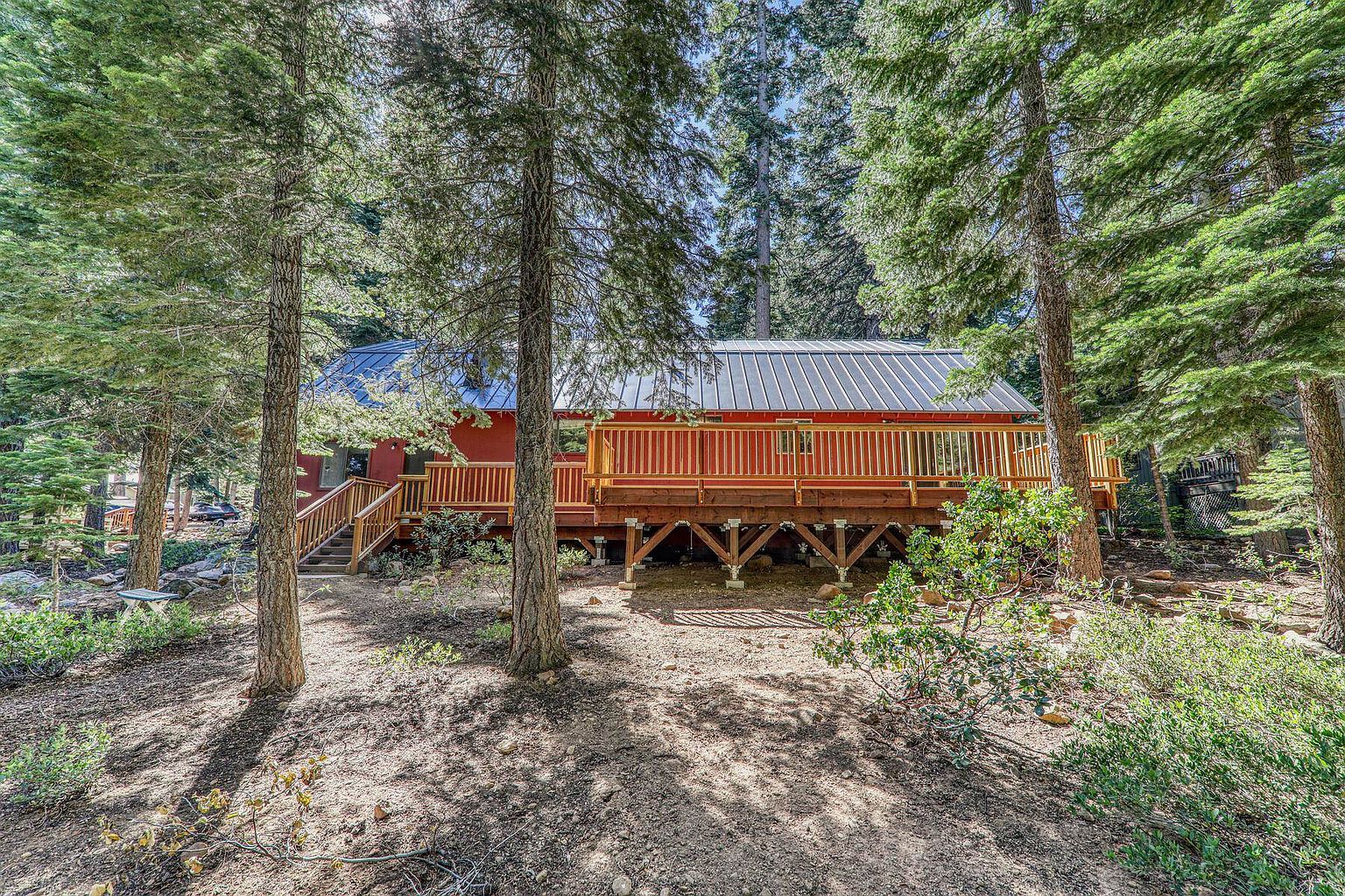 The image showcases a charming red house with a spacious wooden deck, elevated and supported by wooden beams. The deck features a wooden railing and is accessible via wooden stairs. The house is nestled among tall trees, creating a secluded and natural setting.