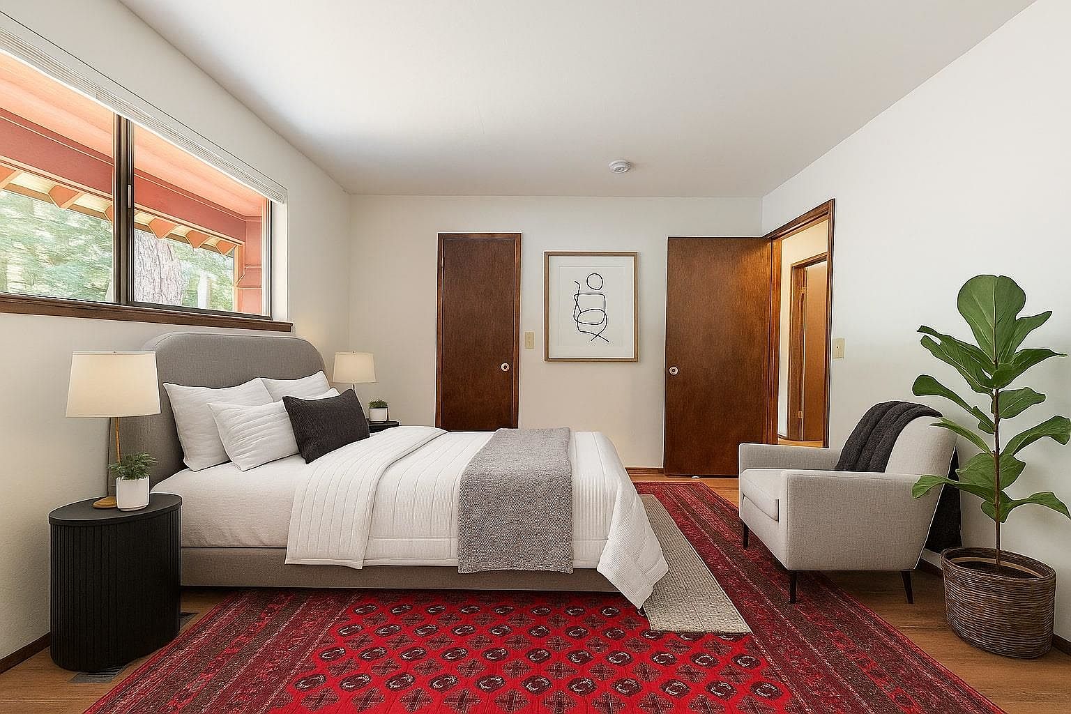 This is a well-staged primary bedroom featuring a gray upholstered bed with white linens and a gray throw. A red patterned rug adds warmth and color to the hardwood floors. The room includes a modern side table with a lamp, an armchair, and a potted fiddle-leaf fig tree, creating a comfortable and inviting atmosphere.