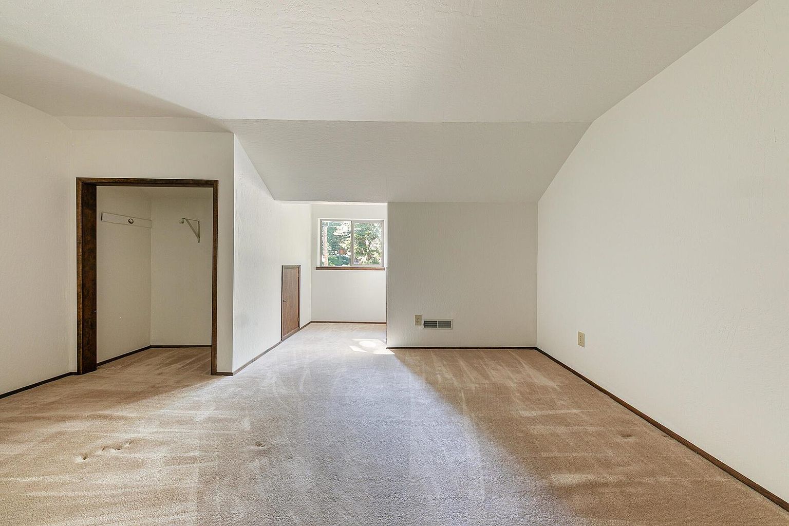 The image showcases a bright, empty bedroom with neutral beige carpeting and white walls. A closet with dark wood trim is visible to the left, and a window provides natural light in the back. The room appears clean and well-maintained, offering a blank canvas for potential buyers.