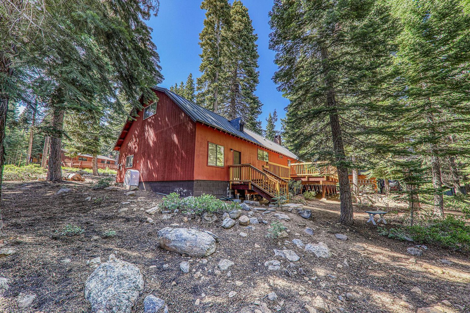 This image showcases the side of a charming, rustic cabin nestled among tall pine trees. The cabin features a reddish-orange exterior with a dark roof, a wooden deck with stairs, and a stone foundation. The surrounding landscape includes a mix of rocks, shrubs, and natural ground cover, creating a serene and secluded atmosphere.
