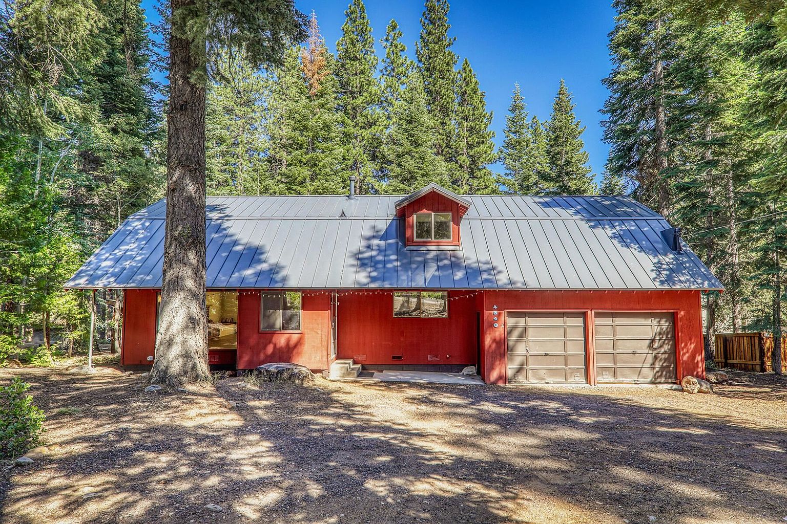 The image showcases the front exterior of a charming red house with a gray metal roof, nestled among tall evergreen trees. The house features a two-car garage, a dormer window, and a well-maintained yard, creating a cozy and inviting atmosphere. The natural lighting and surrounding greenery enhance the property's appeal.