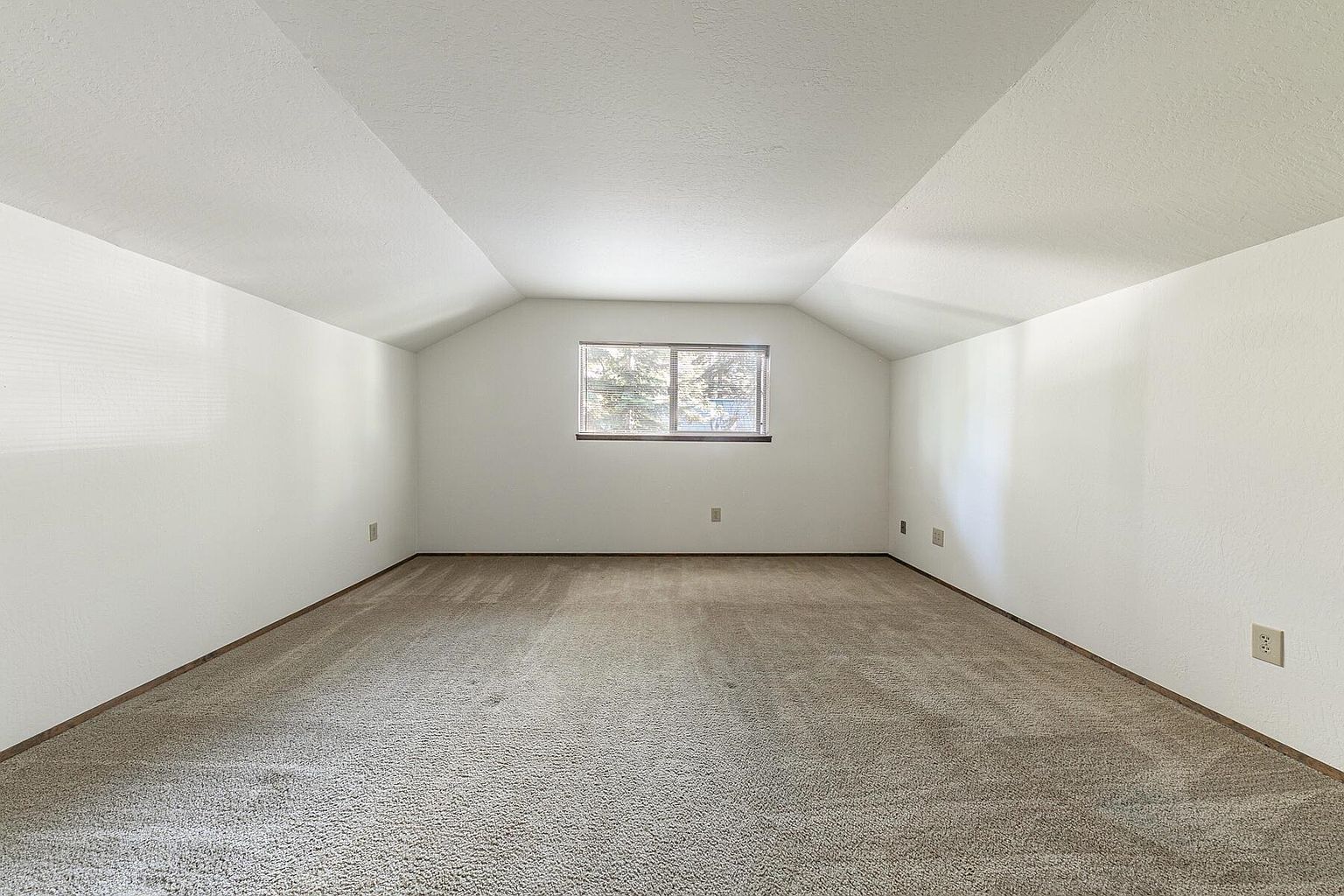This is an interior shot of an attic space. The room features beige carpeting, white walls, and a single window providing natural light. The ceiling slopes inward, creating a unique architectural detail, and the space appears clean and well-maintained, suggesting potential for use as a bonus room or storage area.