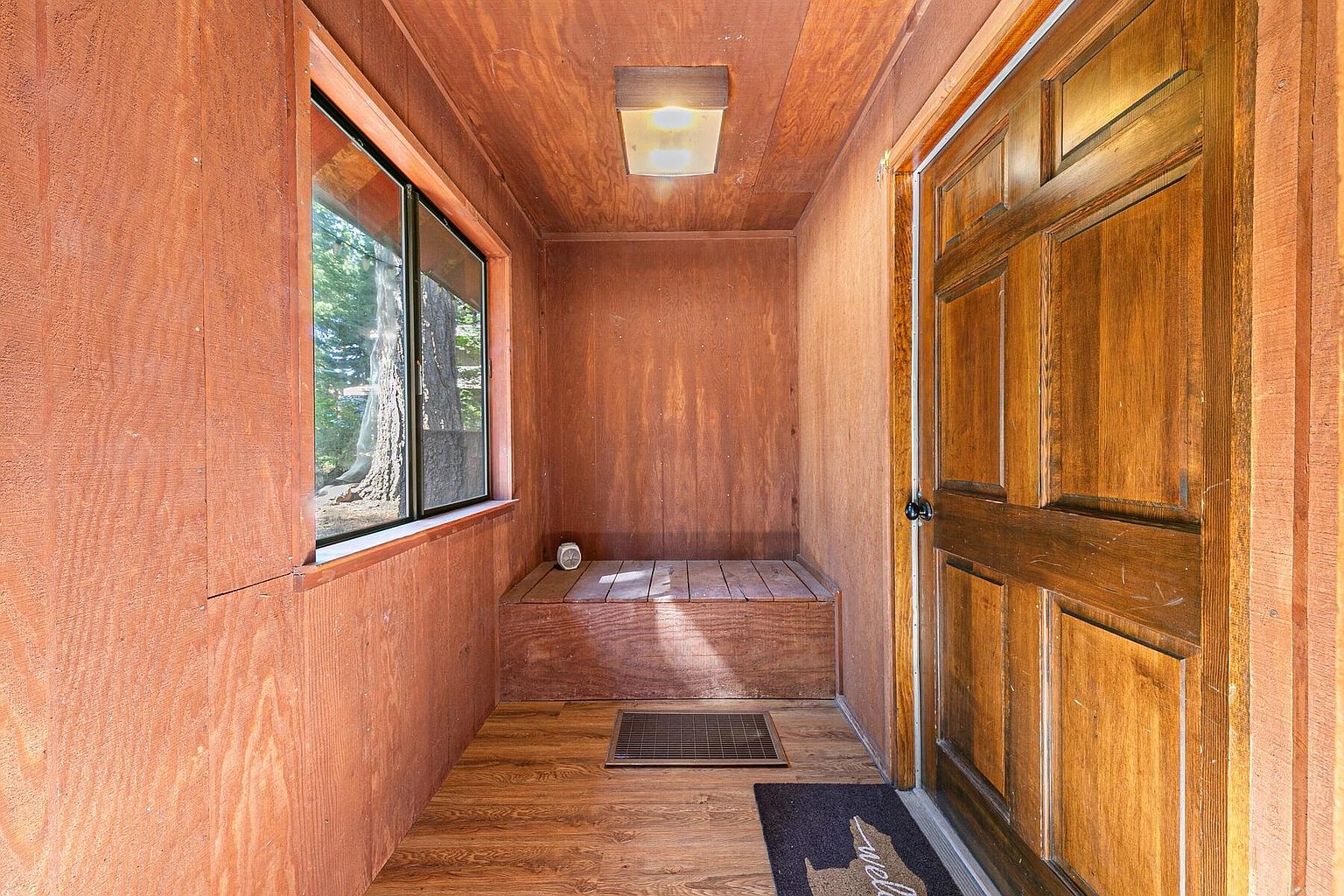This is an interior view of an entryway featuring wood paneling on the walls, ceiling, and a built-in bench. A window on the left provides natural light, while a light fixture illuminates the space from above. A wooden door is on the right, and a welcome mat is partially visible on the floor.