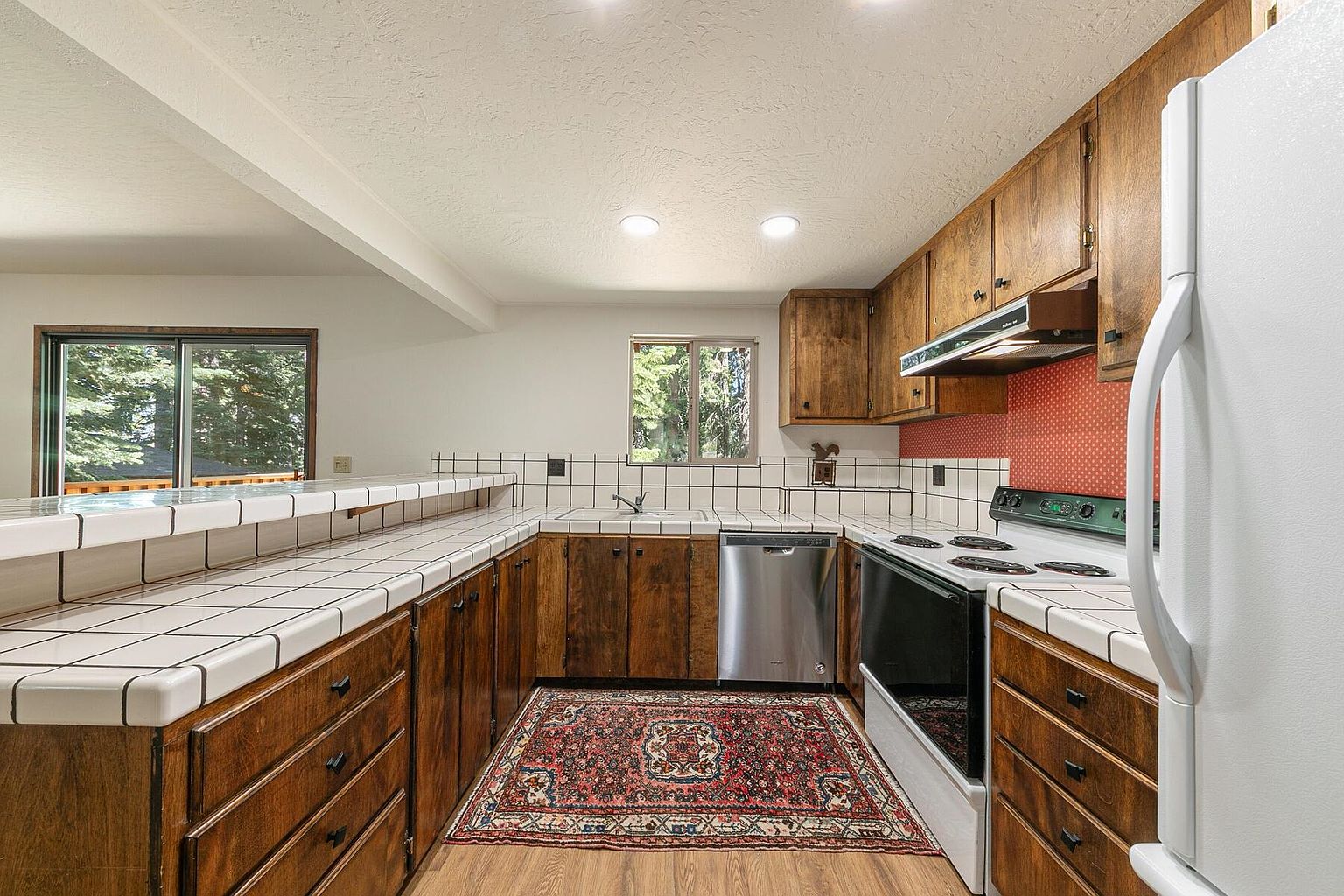This is a kitchen with wood cabinets and white tile countertops. The appliances are stainless steel and white. A colorful rug adds a touch of warmth to the wood floor.