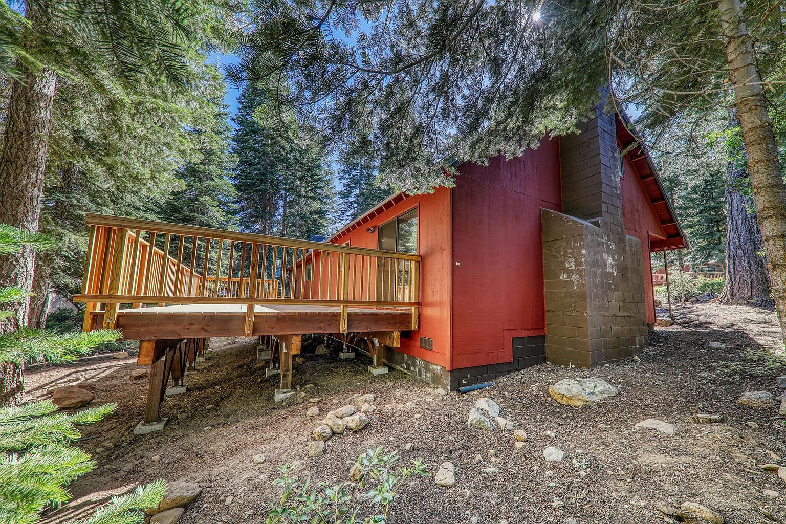 This image showcases the side of a charming red cabin-style home, featuring a wooden deck that extends from the side of the house. The exterior includes a brick chimney and is surrounded by lush greenery and trees, creating a secluded and natural setting. The perspective is from a low angle, emphasizing the height of the house and the surrounding trees.