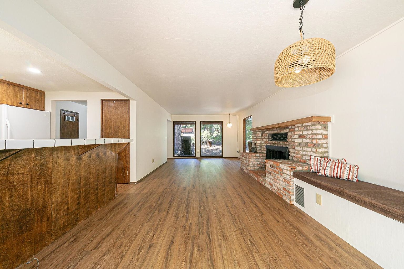This is an interior shot of a living room featuring hardwood floors, a brick fireplace with a built-in bench, and a unique woven pendant light. The room has a warm and inviting feel, with natural light coming in from the doors leading to the outside. The perspective is a wide shot, showcasing the open layout and connection to the kitchen area.