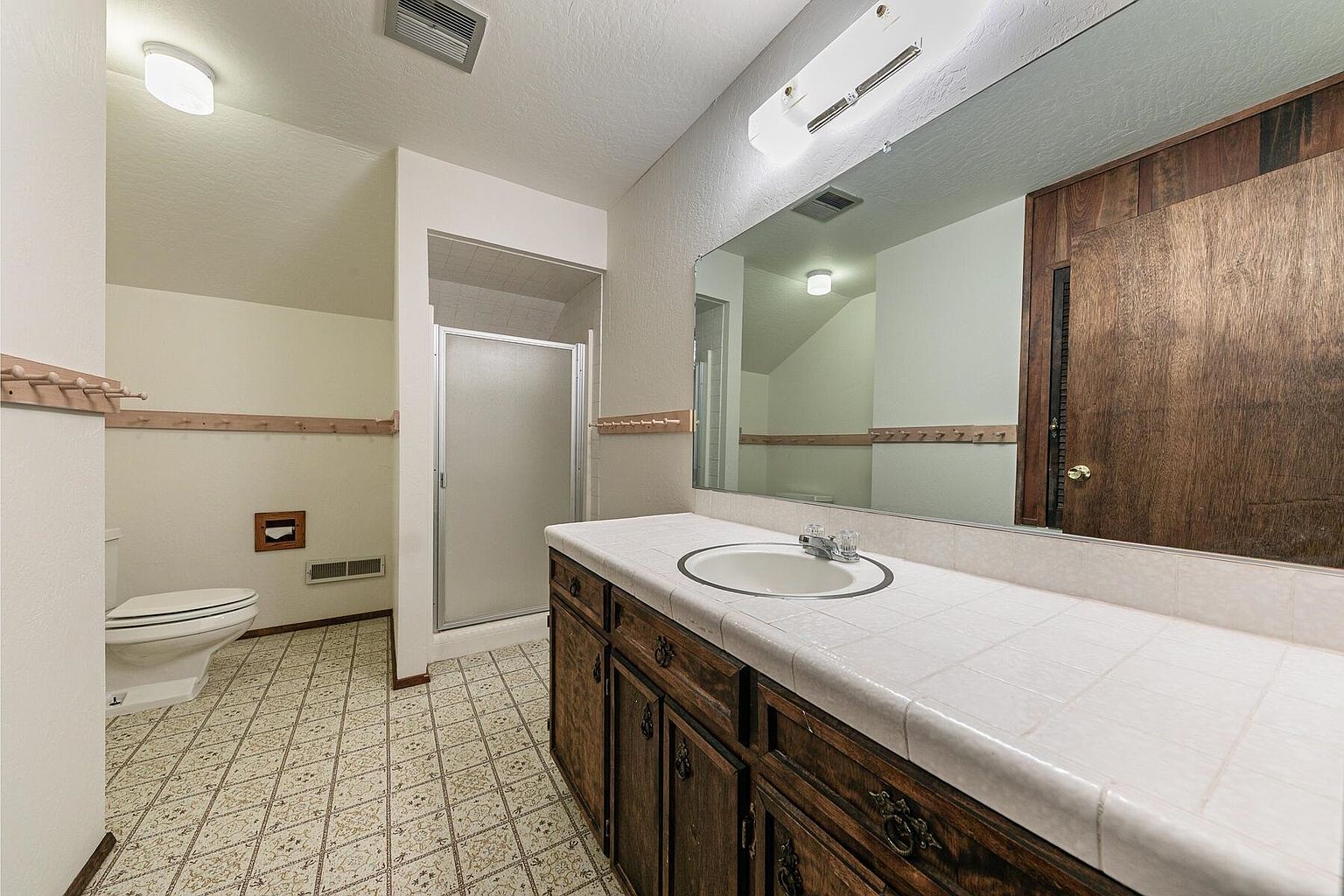 This is a bathroom featuring a tiled floor, a toilet, a shower with a glass door, and a vanity with a white tiled countertop and dark wood cabinets. A large mirror hangs above the vanity, reflecting the bathroom's features and creating a sense of spaciousness. The overall style appears to be dated but clean.