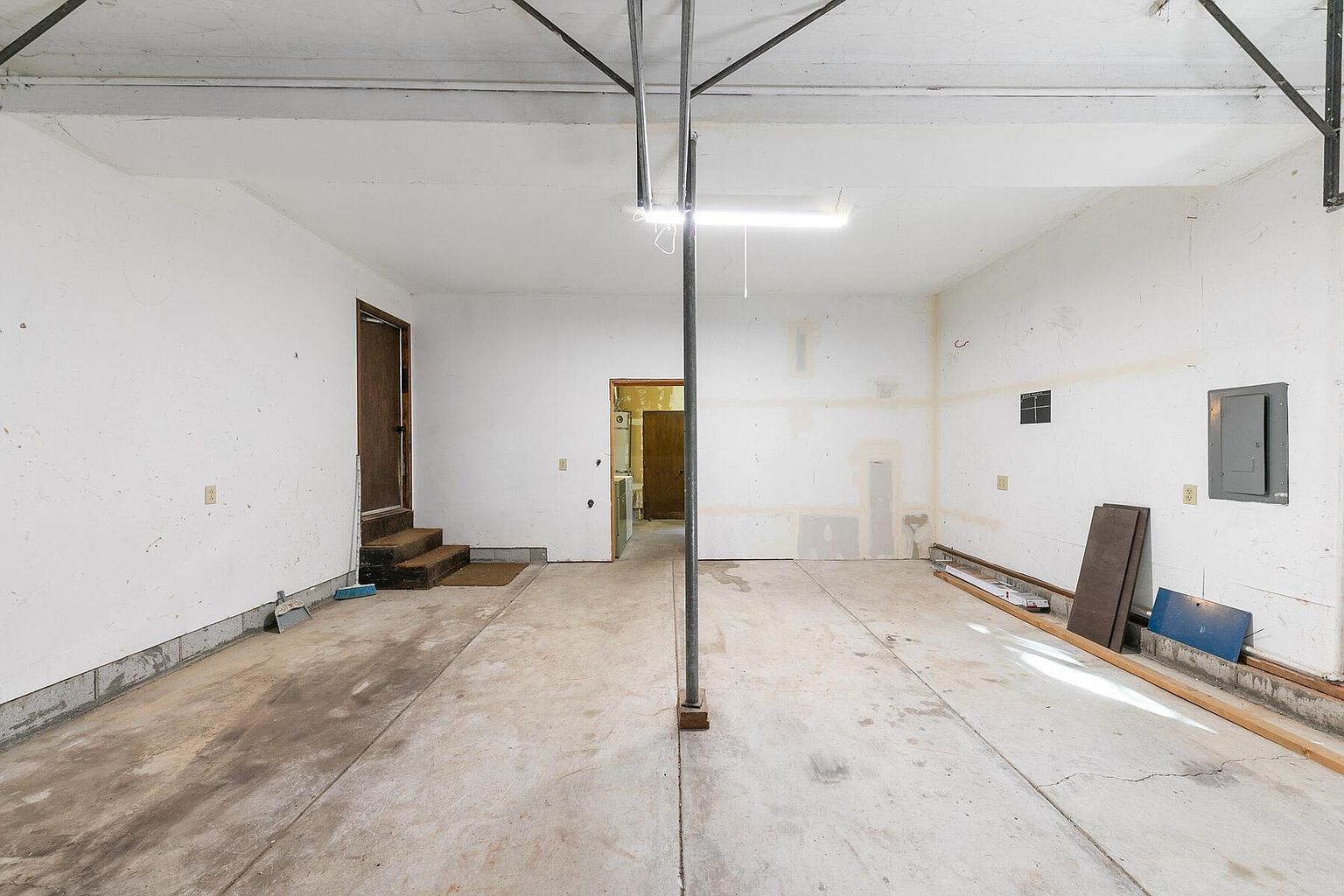 This is an interior shot of a garage. The space is large and open, with a concrete floor and white walls. There is a door leading to another room, and some building materials are stacked against one wall. The overall impression is that the garage is unfinished but has potential.