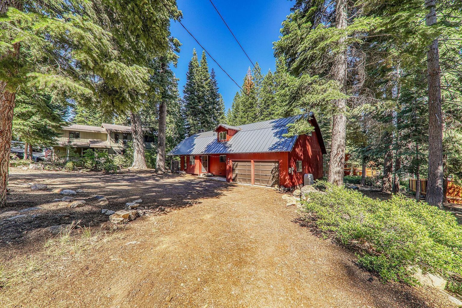 This is a front exterior view of a charming red house with a gray metal roof, nestled among tall pine trees. The house features a two-car garage and a well-maintained gravel driveway. The surrounding landscape includes natural foliage and a sense of privacy, making it an appealing property.