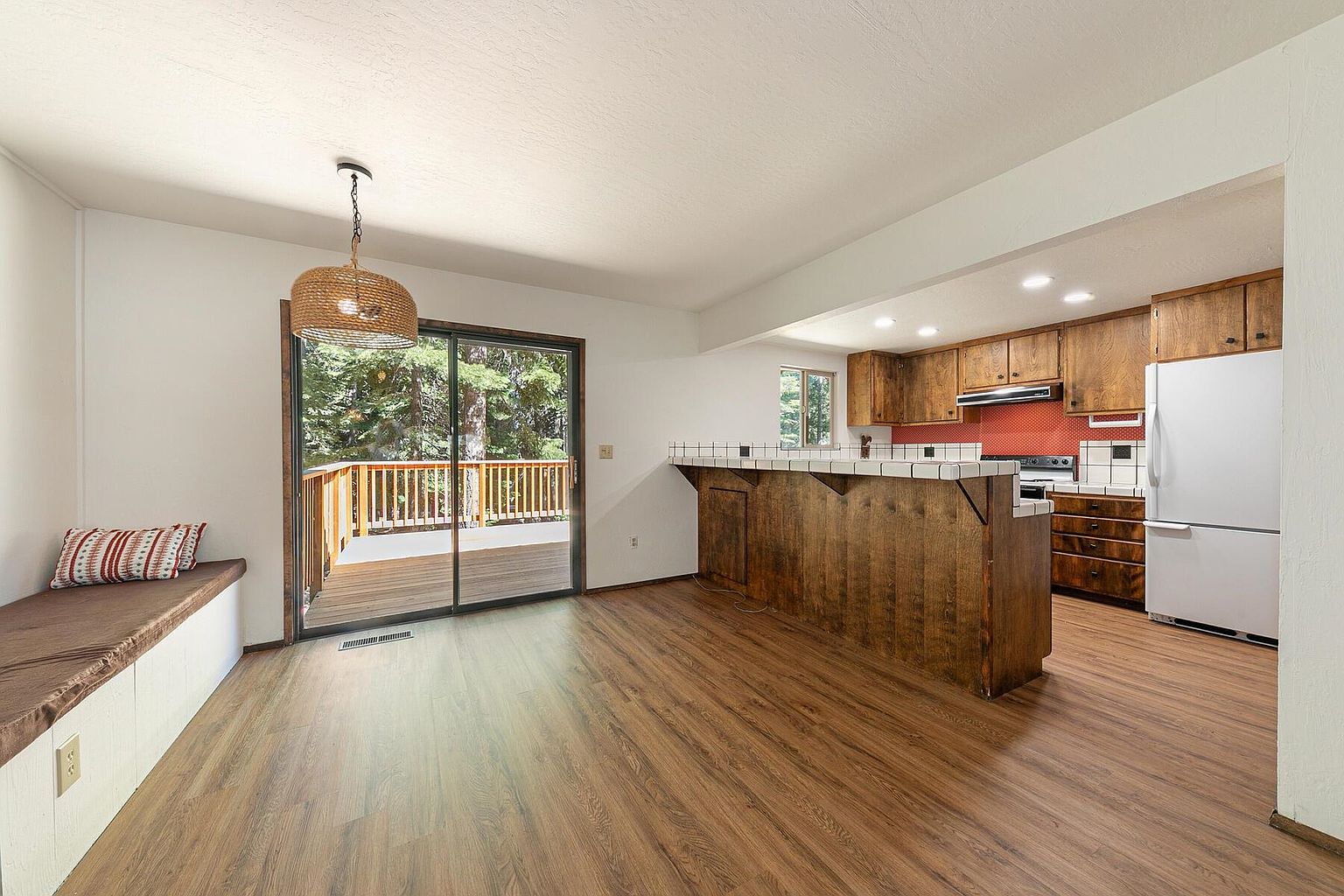 This interior shot showcases a kitchen and dining area with warm wood flooring and cabinetry. A breakfast bar with tiled countertops separates the kitchen from the dining space, which features a built-in bench seat and sliding glass doors leading to a deck. The kitchen is equipped with standard appliances, and the overall style is rustic and cozy.
