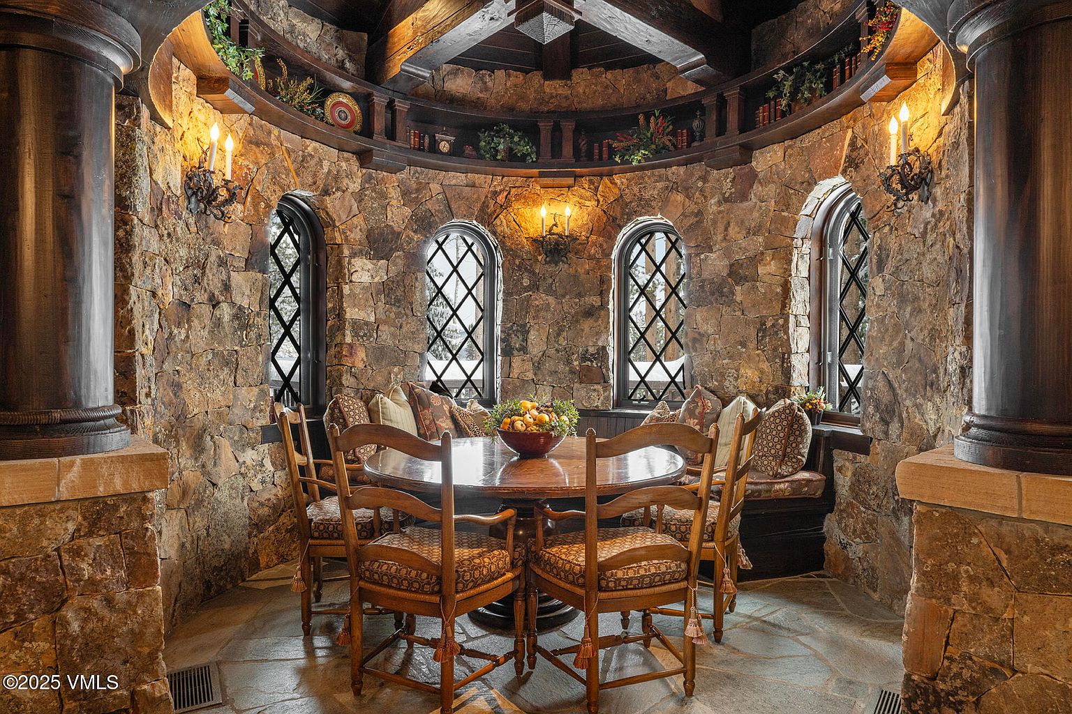 This circular dining area features rustic stone walls and a vaulted ceiling with exposed wooden beams, creating a cozy, lodge-like atmosphere. A round wooden table is surrounded by four upholstered chairs, complemented by a built-in curved bench with patterned cushions nestled against the stone wall. Tall, arched windows with dark grid patterns allow natural light to filter in, while wall-mounted sconces and a circular shelf display add decorative character to the space.