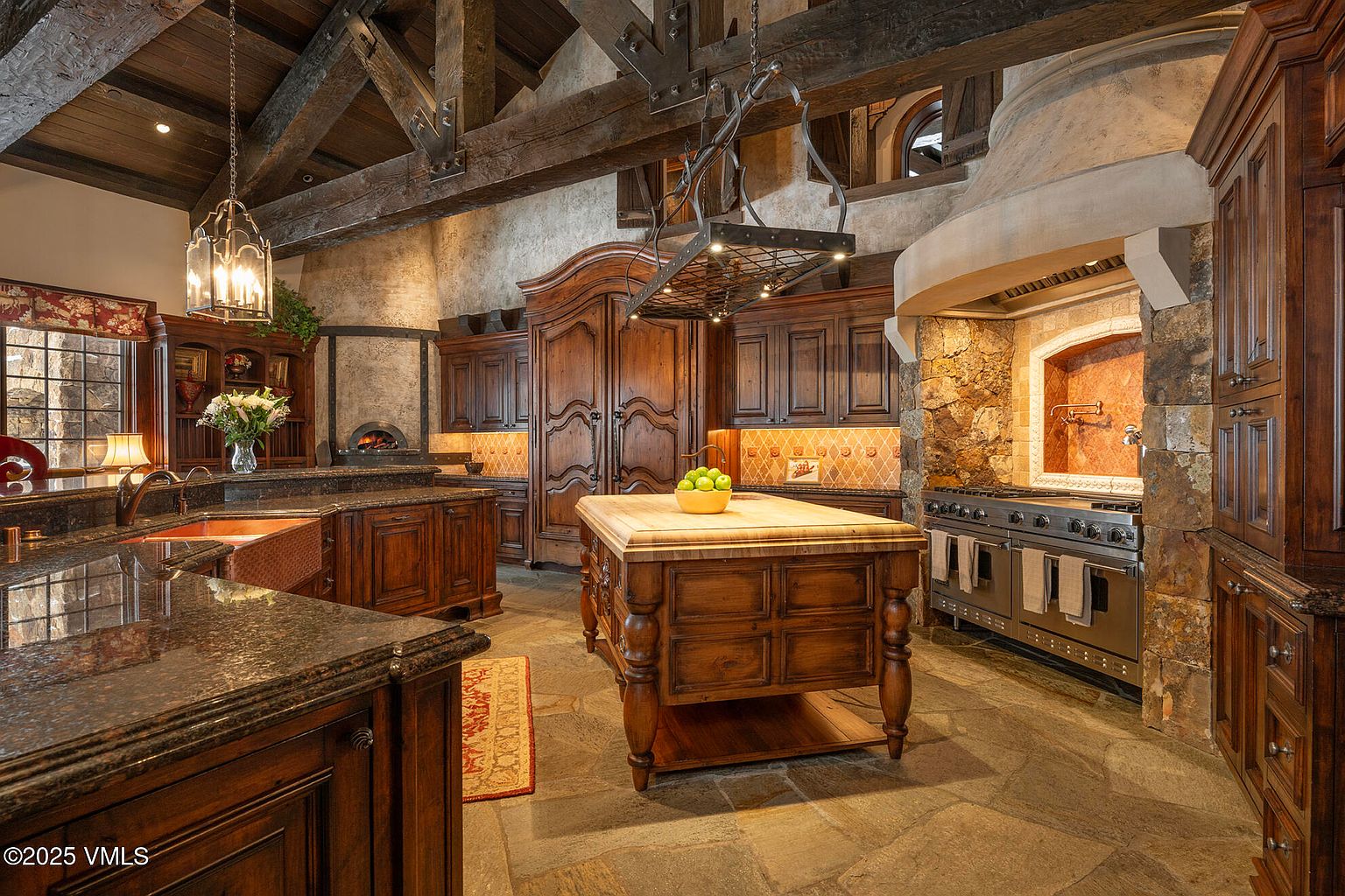 This rustic, high-end kitchen features exposed timber beams, stone flooring, and custom dark wood cabinetry. A central island with a thick wood butcher-block top serves as the focal point, complemented by a professional-grade stainless steel range set within a stone-clad alcove. The space exudes a warm, Old World charm with its intricate iron light fixtures and a built-in pizza oven visible in the background.