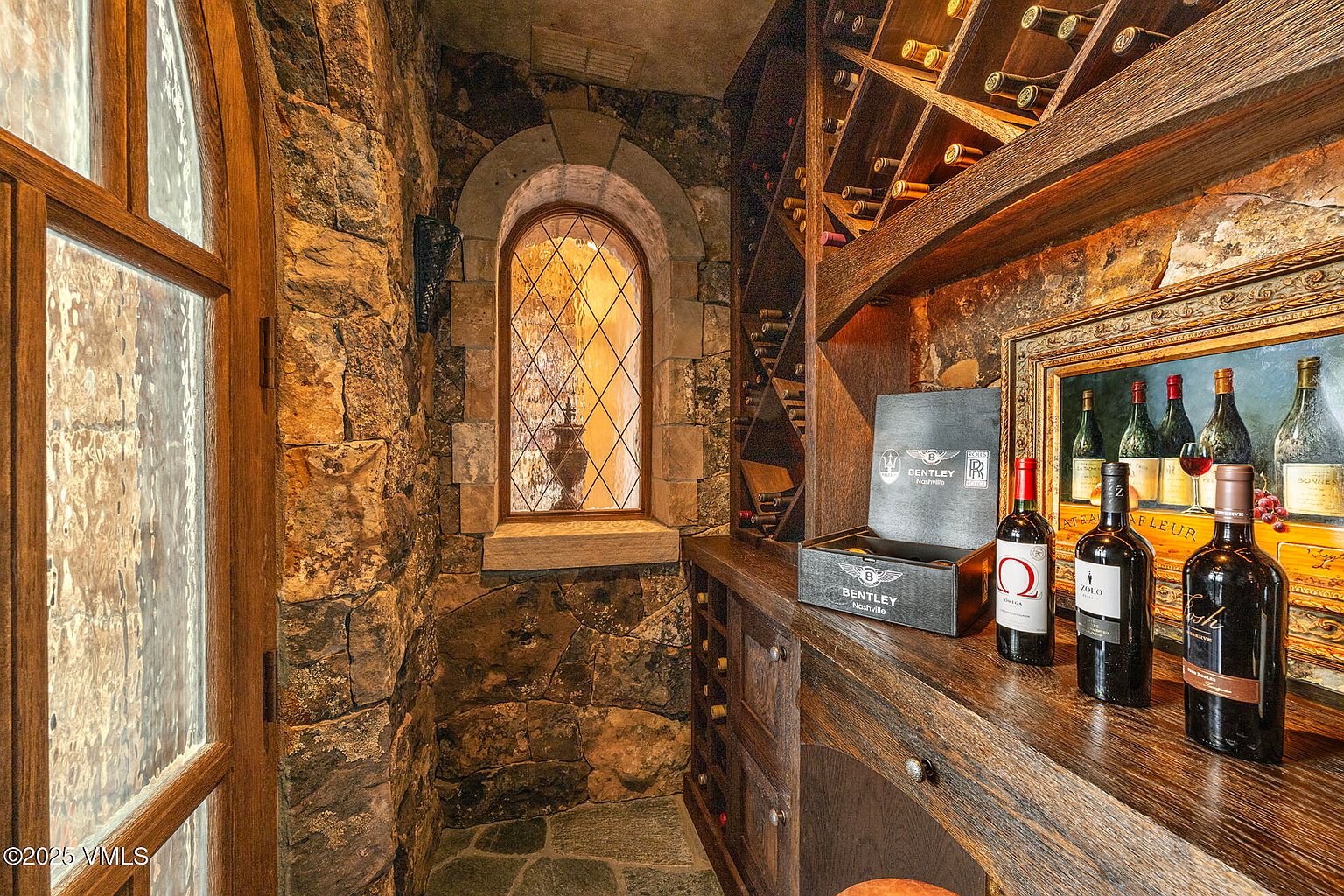 This sophisticated wine cellar features rustic stone walls and custom dark wood wine racking that creates an intimate, old-world atmosphere. A classic arched window with diamond-patterned muntins provides a view into an adjacent space, while a wooden counter displays select wine bottles and a branded Bentley storage box. The stone flooring and warm, ambient lighting enhance the cellar's luxurious and refined aesthetic.