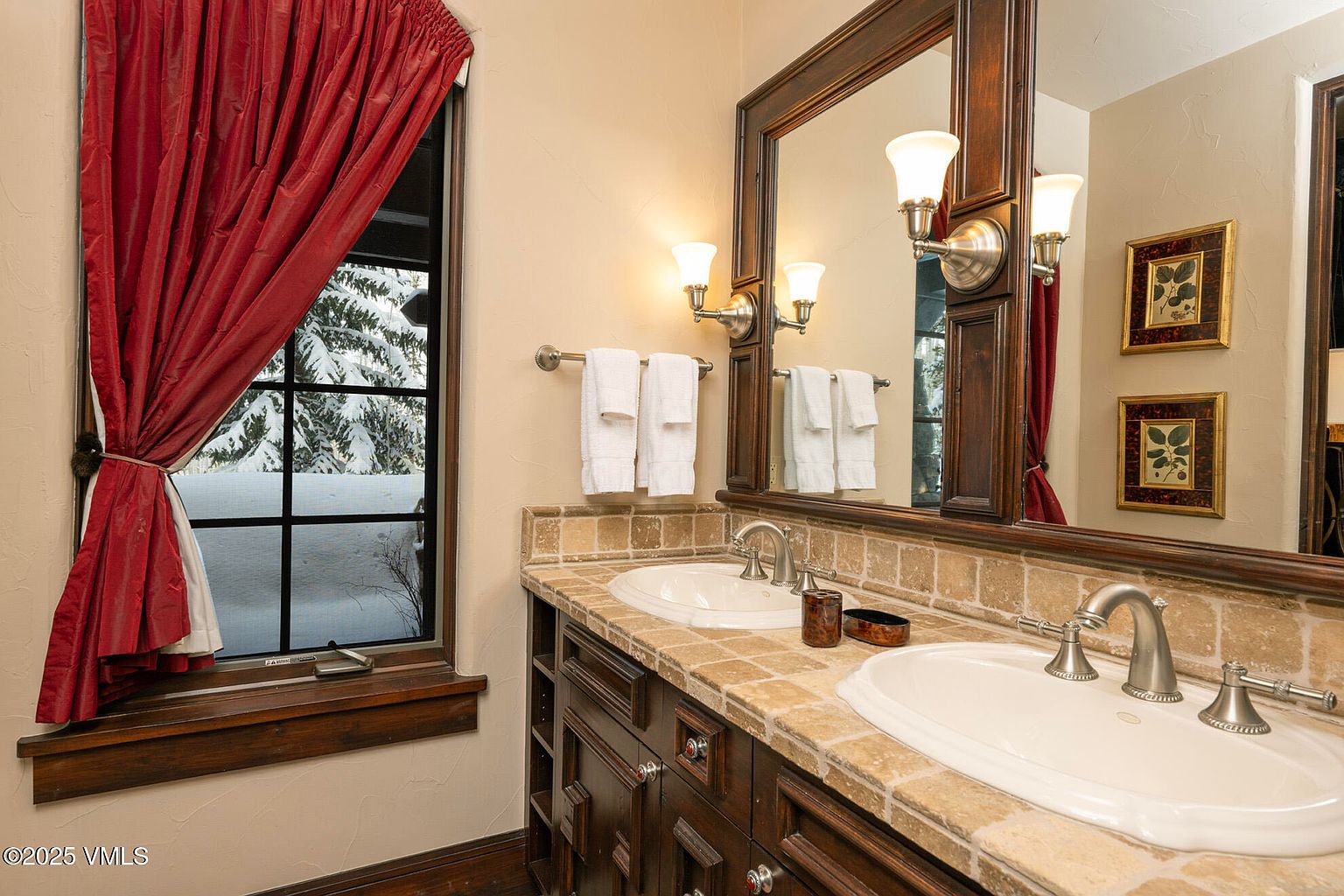This bathroom features a double vanity with a stone countertop and dark wood cabinetry, complemented by a large mirror framed in matching wood. A window with deep red curtains provides a view of a snowy landscape, while wall-mounted light fixtures add warmth to the space. The room is finished with neutral-toned wall tiles and classic plumbing fixtures, creating a cozy and traditional aesthetic.