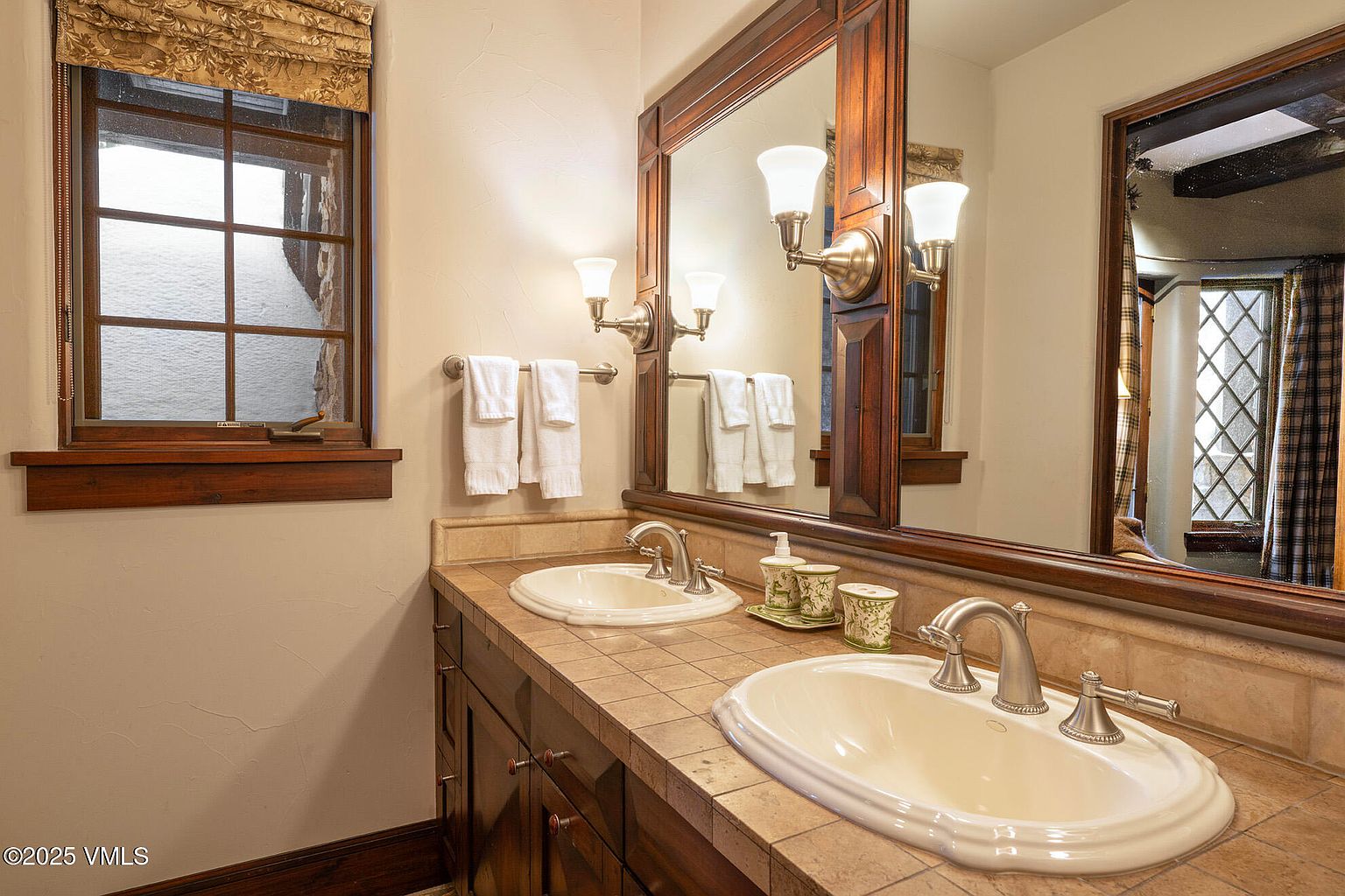 This bathroom features a double vanity with a tiled countertop and two inset sinks, set against a large, wood-framed mirror. The space is accented by warm-toned walls, a window with patterned fabric valance, and decorative light fixtures. The overall aesthetic is traditional and cozy, with a focus on functional, classic design elements.