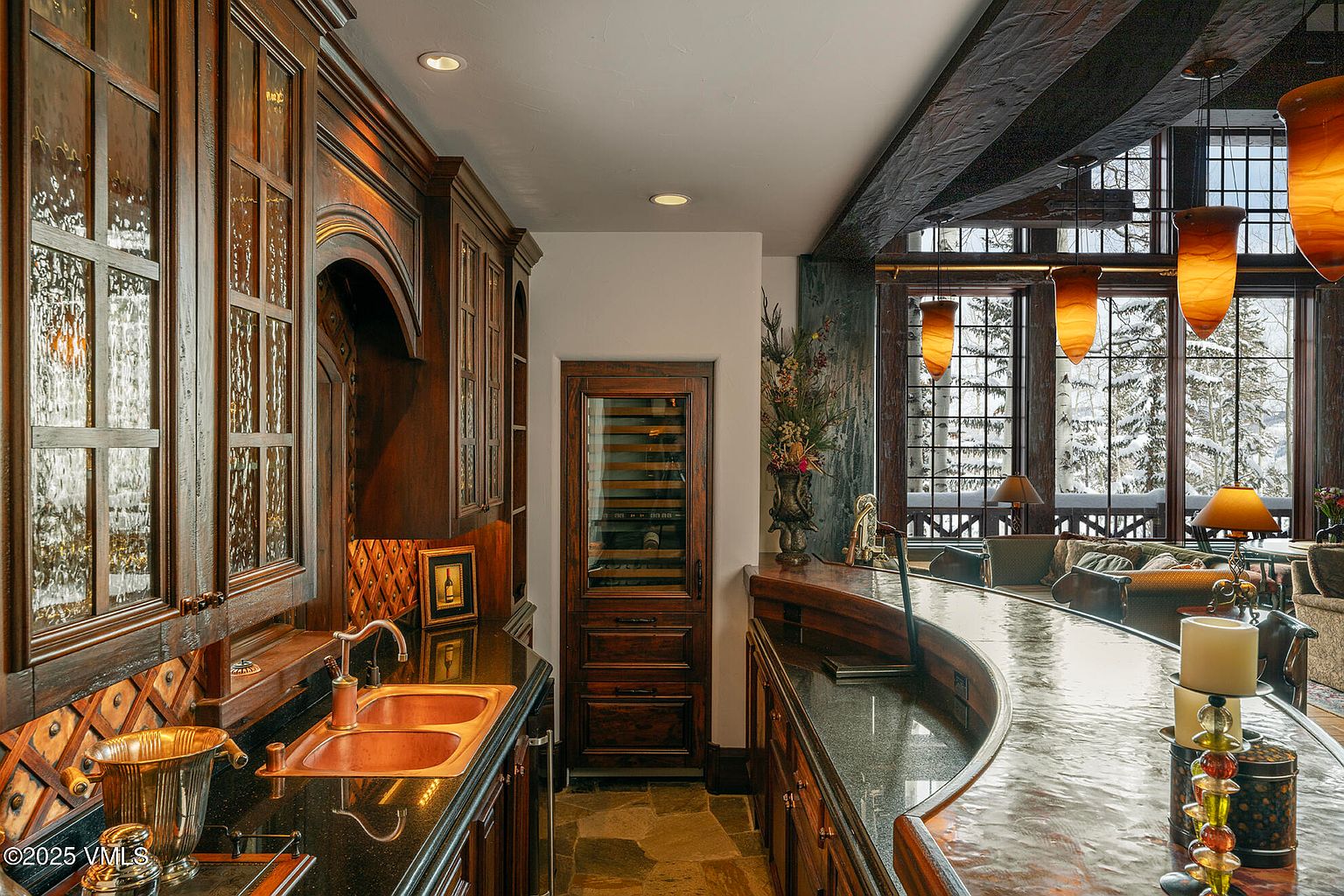 This sophisticated wet bar area features rich, dark wood cabinetry, a polished stone countertop, and a double-basin copper sink. A built-in wine refrigerator is integrated into the cabinetry, while warm pendant lighting hangs above a curved bar counter that overlooks a cozy living space with large windows showcasing a snowy landscape. The overall aesthetic is rustic and luxurious, emphasizing high-end craftsmanship and a warm, inviting atmosphere.