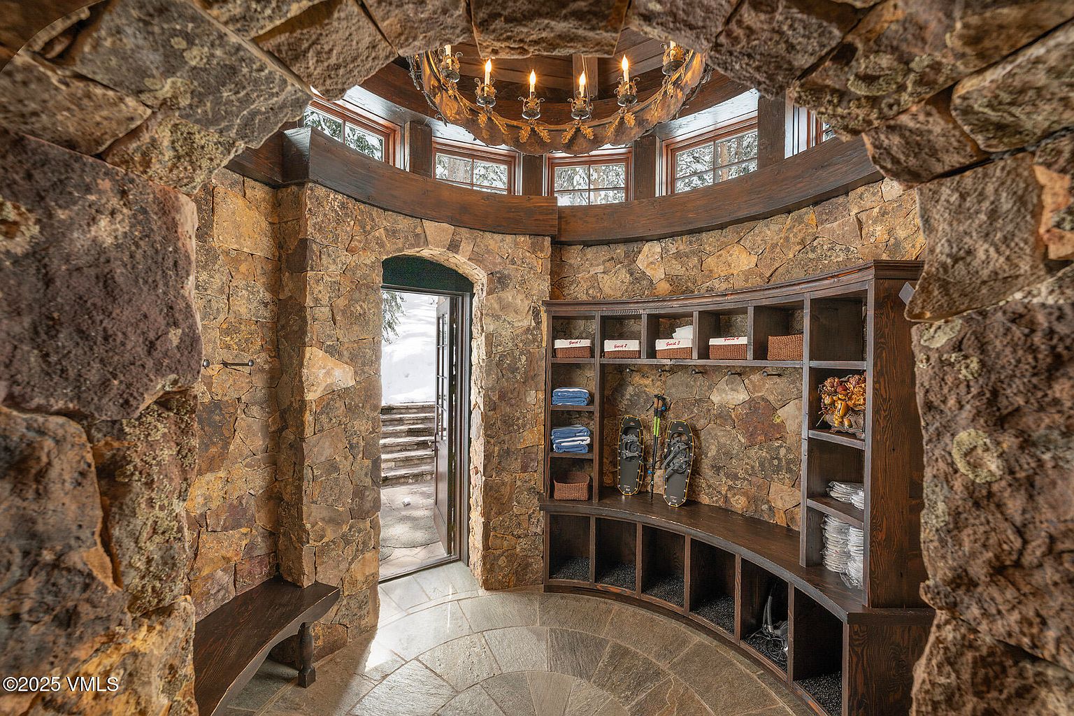 This rustic entryway features dramatic, curved stone walls and a circular layout, creating a grand, fortress-like atmosphere. A custom-built, curved wooden storage unit provides organized cubbies and shelving for gear, while a large, ornate chandelier hangs from the center of the vaulted ceiling. The space opens directly to an outdoor stone staircase, blending indoor storage functionality with a rugged, mountain-inspired aesthetic.