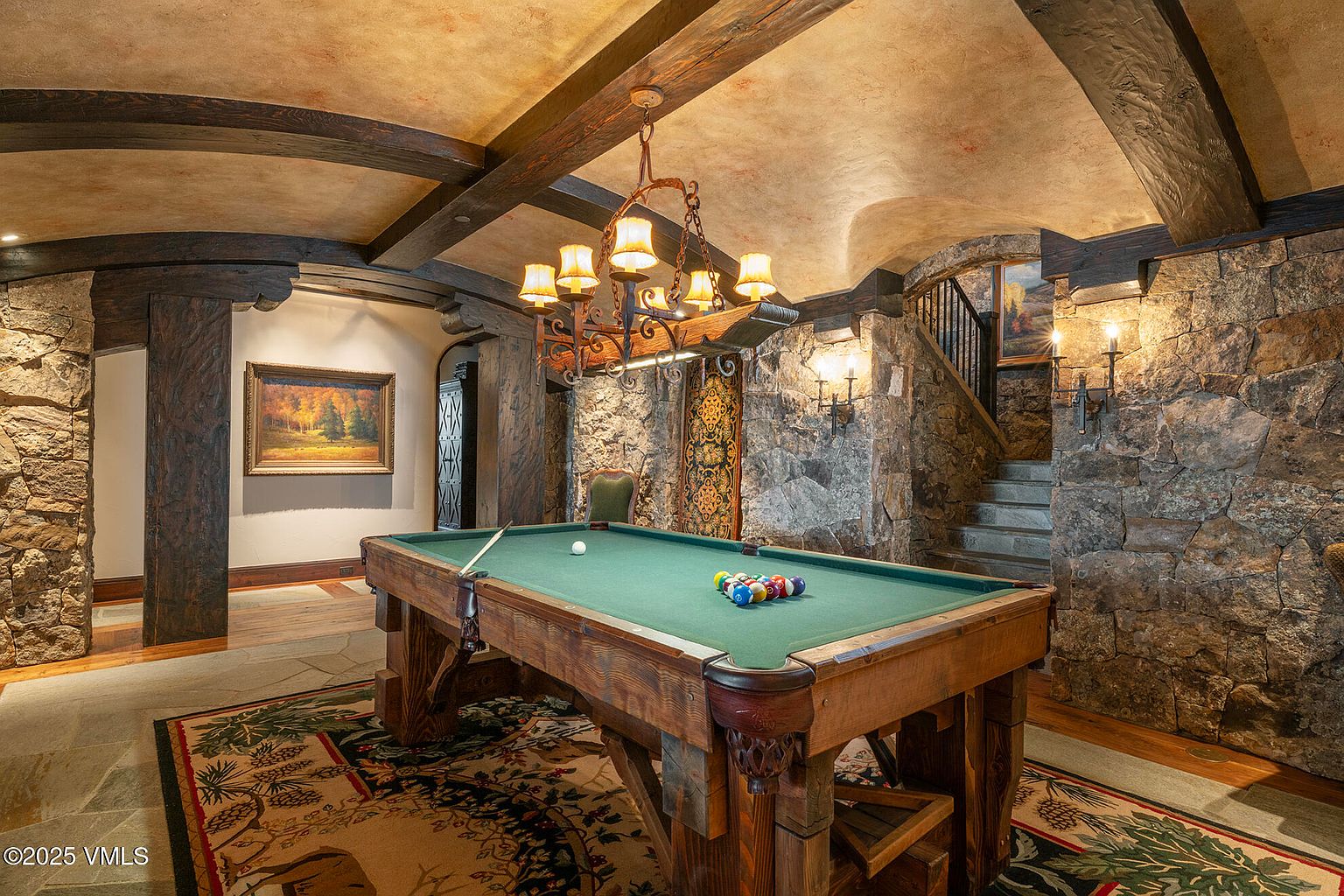 This rustic, lodge-style game room features a prominent pool table as the centerpiece, set beneath a decorative wrought-iron chandelier. The space is defined by rugged stone walls, exposed dark wood ceiling beams, and a barrel-vaulted ceiling, creating an intimate, tavern-like atmosphere. A staircase with a wrought-iron railing leads to an upper level, while framed artwork and a decorative wall hanging add character to the stone-clad walls.