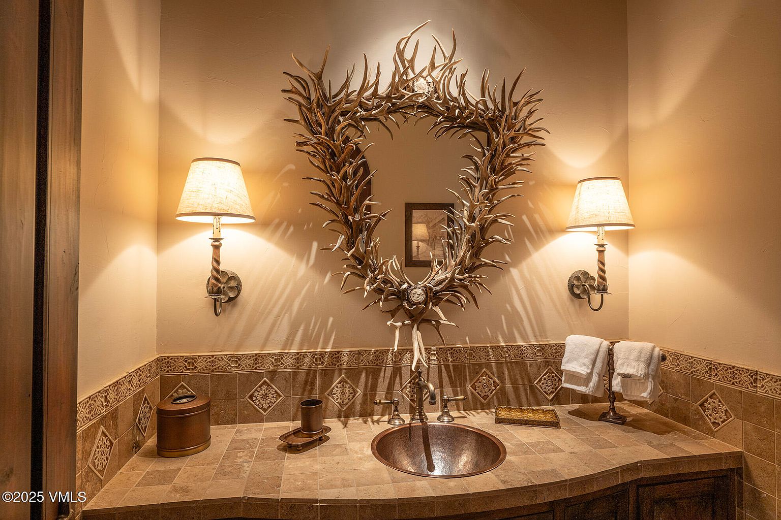 This rustic powder room features a unique mirror framed by natural antlers, serving as the focal point above a copper vessel sink. The vanity is topped with earth-toned stone tiles that extend to the backsplash, complemented by matching wall sconces with fabric shades. The warm lighting and textured wall finish create an inviting, lodge-inspired atmosphere.